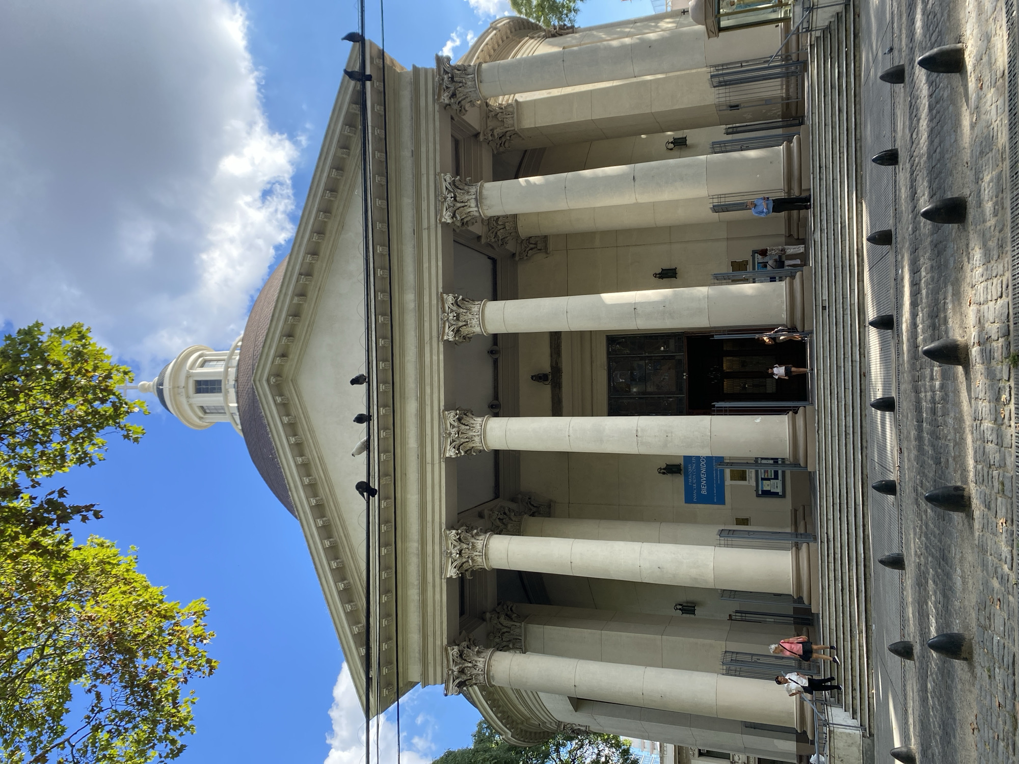 The image depicts the Church of the Immaculate Conception in Buenos Aires, Argentina. The building has a neoclassical façade with tall Corinthian columns supporting a triangular pediment. A dome rises behind the pediment. Several pigeons are perched on the top of the building and on the power lines in front of it. People are walking up and down the steps leading to the entrance. A blue sign with white writing is visible near the entrance. A brick sidewalk with black bollards runs along the side of the building. Trees with green leaves are visible on the left side of the image, contrasting with the blue sky and white clouds.