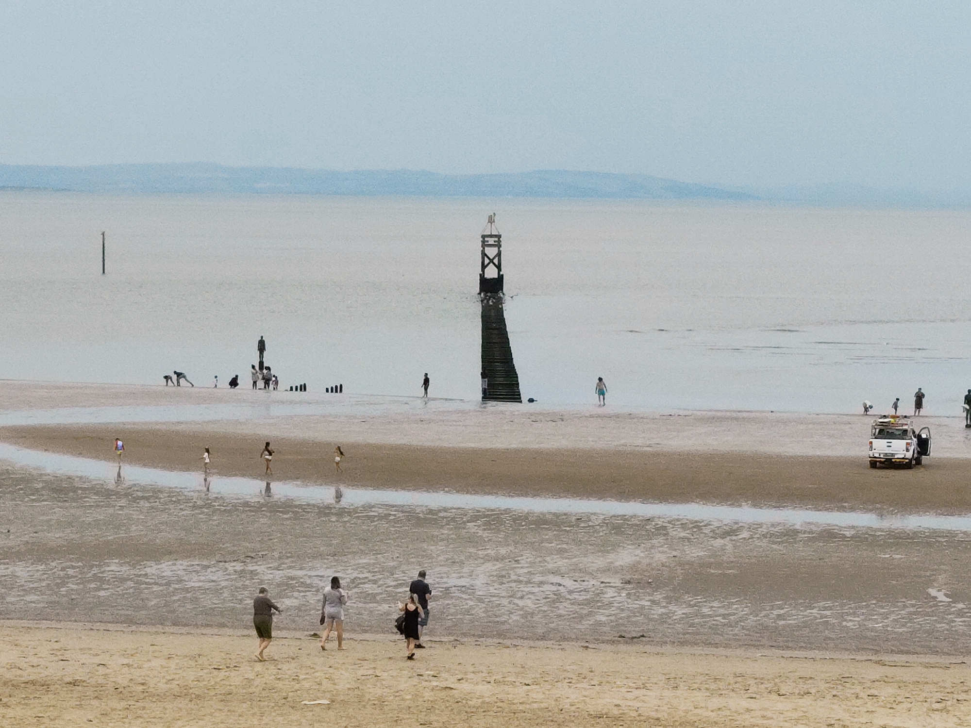 The image captures a wide, sandy beach at low tide, likely Crosby Beach in the United Kingdom, given the presence of the wooden structures known as "Another Place" sculptures, one of which is visible in the middle distance. The sky is overcast and pale blue, with a hazy horizon suggesting distant landforms. The water is calm and shallow, reflecting the muted light.
In the foreground, several individuals are walking on the wet sand. A man and two women are walking towards the viewer, while another person is walking away. Further into the mid-ground, a diverse group of people are spread across the exposed seabed. Some are walking, while others appear to be playing or exploring. A small, white utility vehicle, possibly a lifeguard or beach patrol vehicle, is parked on the sand to the right of the frame, with its driver's door open.
Several of the "Another Place" sculptures, which consist of cast-iron figures of a man, are scattered across the beach and in the shallow water. The most prominent sculpture in the image features a tall wooden structure with steps leading up to it, topped with a cross-like fixture. Other wooden posts, remnants of a former jetty or marker, are also visible in the distance. The overall scene conveys a sense of quiet activity on a beach at low tide under a diffused sky.