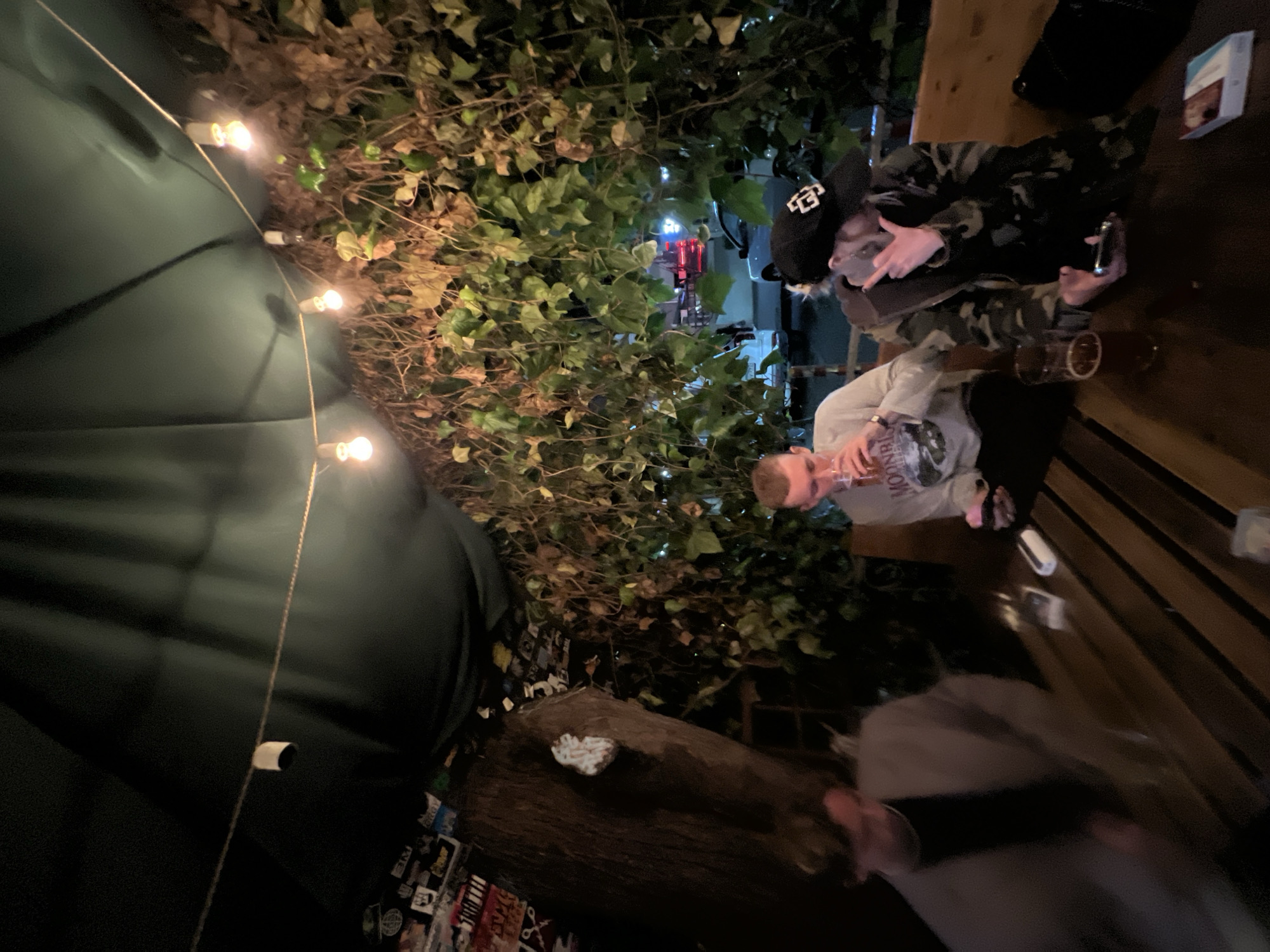 Two people are sitting at a table outside, engaged in conversation. One individual, wearing a camouflage jacket and a black hat with a white logo, holds a silver object in their hand. Their companion is dressed in a light-colored sweatshirt. A glass of dark liquid sits on the table between them. String lights illuminate the area overhead, draped around greenery. The location, based on the presence of Cyrillic script and context clues, is likely in a public outdoor area in the city of Sochi, Russia. The scene depicts a casual, social interaction.