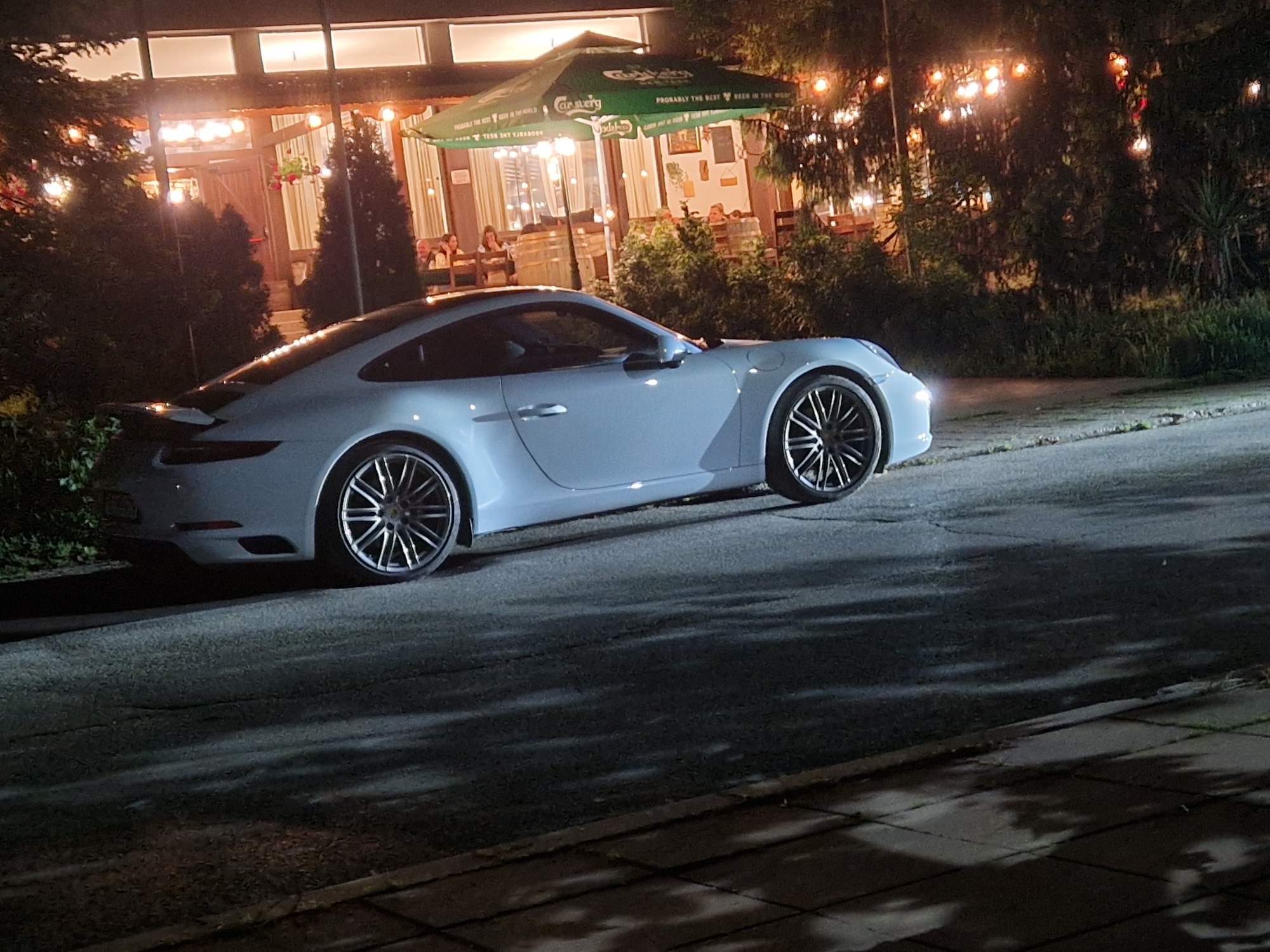 The scene depicts a nighttime urban environment in Razgrad, Bulgaria. A light-colored Porsche 911, with its headlights active, is parked on an asphalt street, oriented towards the right side of the frame. The vehicle is equipped with dark, multi-spoke wheels.
To the left and behind the vehicle, a restaurant or cafe building is prominently illuminated. Its facade features large windows and visible wooden architectural elements. The area around the building is brightly lit by internal lights and numerous warm-toned string lights draped over trees and along the structure.
An outdoor seating area is visible, partially covered by a large green Carlsberg umbrella, which displays the text "PROBABLY THE BEST BEER IN THE WORLD." Several individuals are present, seated at tables both inside and outside the establishment, engaged in dining or social interaction. Ornamental evergreen trees and various bushes are planted around the restaurant's entrance and along the street.
The asphalt street surface exhibits minor cracking. A concrete-paved sidewalk runs parallel to the street on the right, displaying distinct shadows cast by unseen overhead foliage. The overall lighting is dim, concentrated around the restaurant and car, indicative of a nighttime setting.