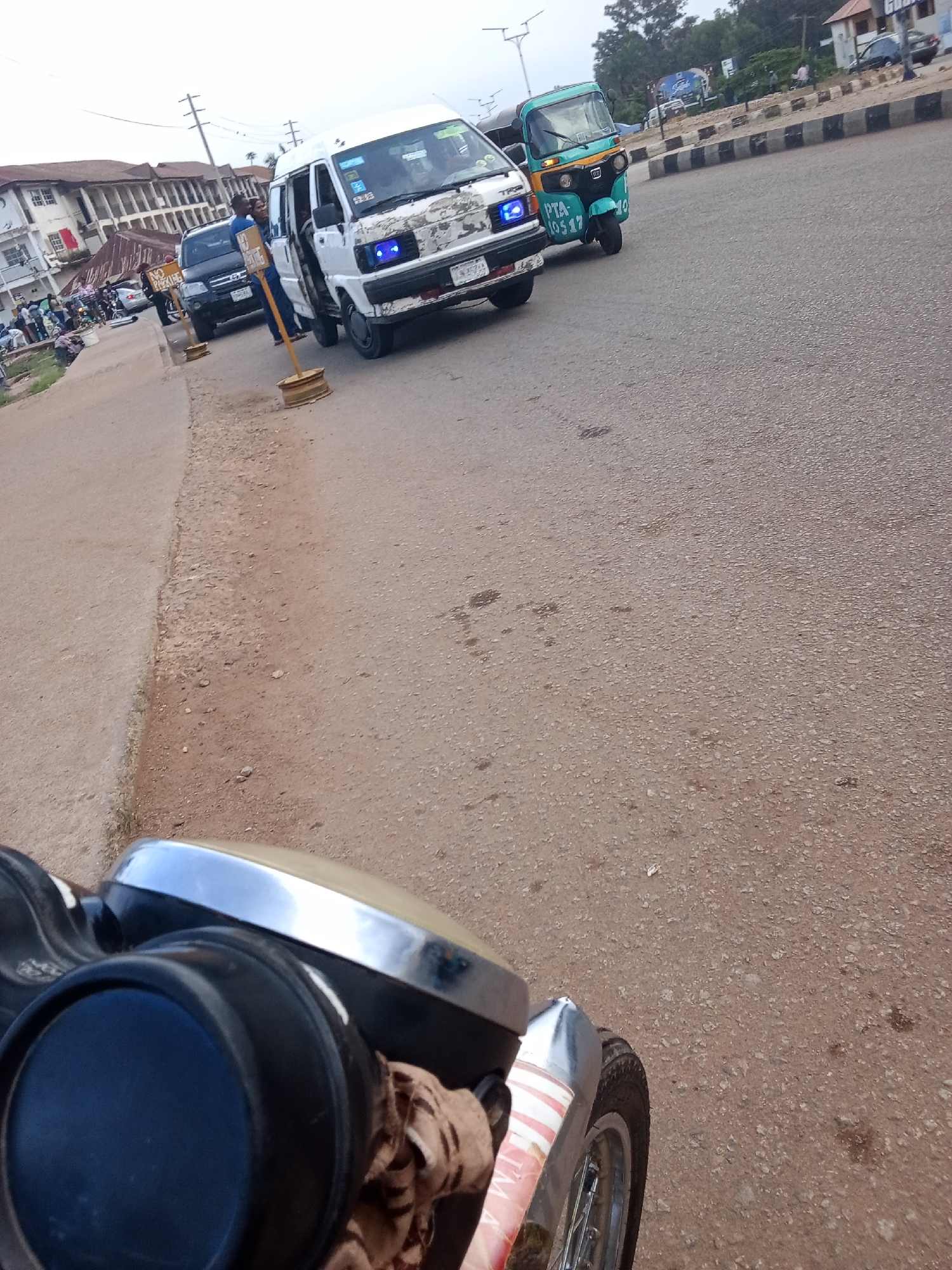 The image depicts a street scene in Gura Top, Nigeria, with a focus on vehicles and the surrounding urban environment. In the foreground, a portion of a motorcycle is visible, including its black handlebar, a chrome headlight housing, and a tire with a red and white striped mudguard. The motorcycle is positioned on an asphalt road, which shows signs of wear and tear, with scattered gravel and dirt.
Further down the road, a white minibus with blue headlights is parked, and a man in a blue shirt stands beside it, seemingly interacting with a yellow signpost. Another vehicle, a black SUV, is parked behind the minibus. To the right of the minibus, a green tricycle, a common mode of transport in Nigeria, is visible. This tricycle has "PTA-10517" written on its side. The background shows buildings with traditional architecture and power lines, suggesting a residential or commercial area within the city. Pedestrians are scattered along the roadside, indicating a lively, active environment. The sky is overcast.