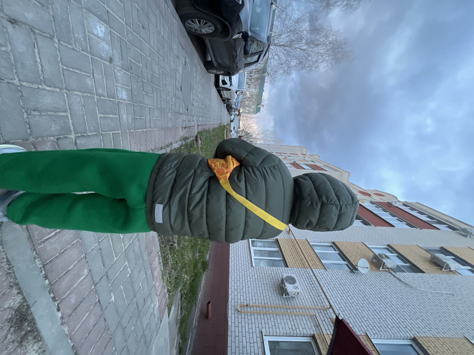A person in green pants and an olive green hooded puffer jacket stands on a brick sidewalk in Usman, Russia. The individual has an orange and yellow patterned item with a bright yellow strap across their back. To the left of the person, cars are parked along the street. To the right, a light brick building is visible. The sky is overcast.
