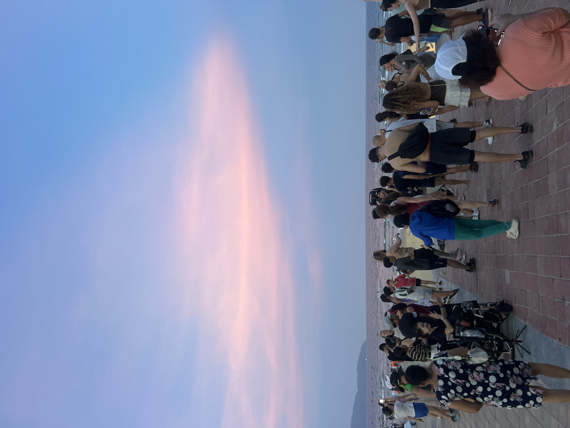 At sunset, a crowd of people gather on a brick walkway by the ocean. The sky is a gradient of soft blues and pinks, with a distinct cloud formation streaked across the horizon. The ocean is calm, with gentle waves lapping at the shore. In the foreground, people are standing and interacting, some appear to be talking, while others are looking out at the sea or taking pictures with their phones. One woman is seated in a wheelchair and appears to be waving. Several people are wearing casual beach attire, such as shorts and t-shirts. The overall atmosphere is relaxed and festive, suggesting a social gathering or event.