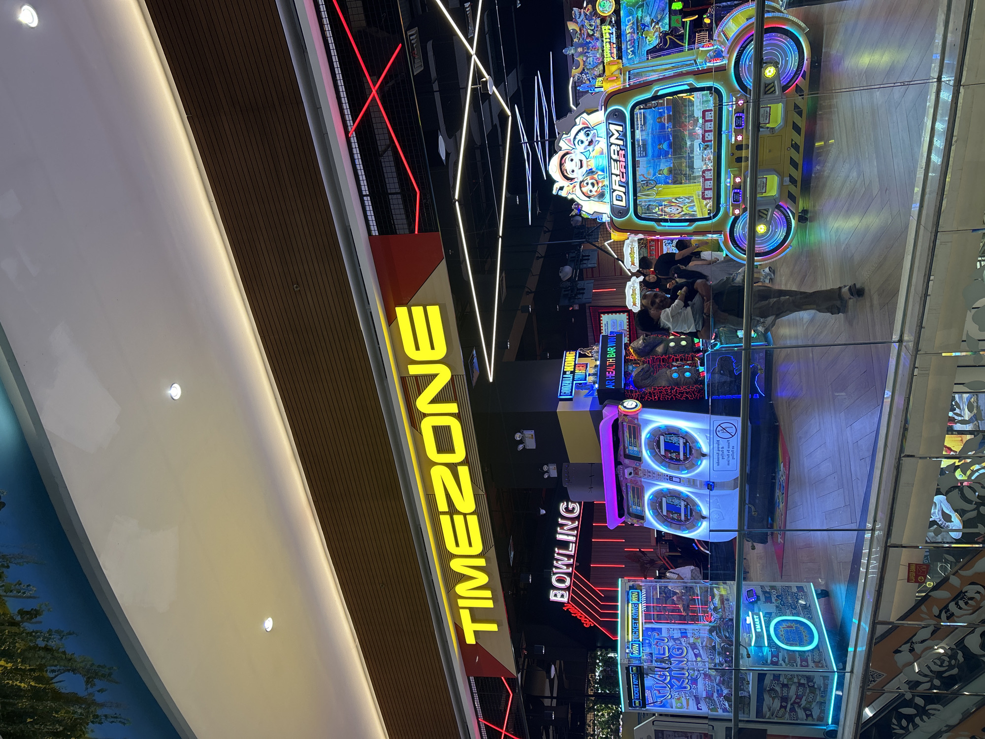 The image shows the interior of a Timezone amusement center, likely located in a shopping mall in Makassar, Indonesia. The foreground features a glass railing and a portion of an escalator, indicating the perspective is from a higher level. Dominating the scene are various arcade machines, illuminated with bright neon lights. A prominent yellow and blue "DREAM CAR" claw machine is situated in the mid-right, with cartoon characters on its top. To its left, two large, futuristic-looking machines with purple and white accents are visible, possibly for ticket redemption or prize dispensing. Further left, a "TICKET KING" machine is lit up in blue and green. In the background, the large yellow neon sign for "TIMEZONE" is clearly displayed above a "BOWLING" sign with red neon accents. Several people are present, including a man holding a child near the "DREAM CAR" machine and other patrons scattered throughout the arcade. The environment is brightly lit and appears to be a recreational entertainment venue within a commercial complex, characterized by its colorful and electronic displays.