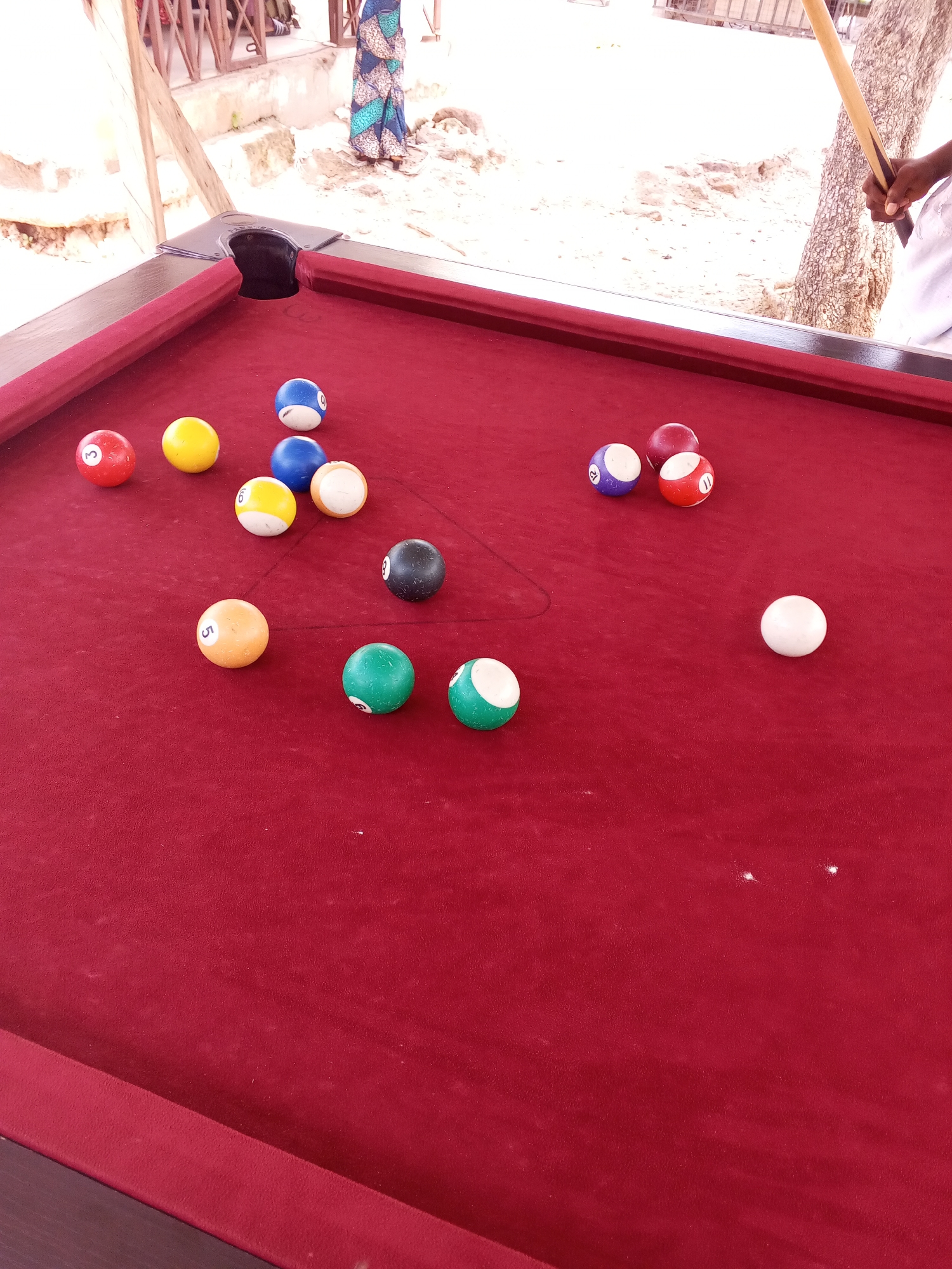 The image shows a close-up view of a red billiard table with several billiard balls. The balls are scattered across the table, with some clustered together and others spread out. The balls are of various colors and numbers, including a red ball with the number 3, a yellow ball, a blue ball with the number 0, a yellow ball with the number 6, a blue ball, a white ball with a partially visible number, a black ball with the number 8, a green ball, a white and green ball, an orange ball with the number 5, a purple ball with the number 12, a red ball with the number 11, and a solid red ball.
In the background, there is a person holding a pool cue, suggesting that a game of billiards is in progress or about to begin. The background also shows a sandy area with some vegetation and structures, hinting at an outdoor or semi-outdoor setting, possibly a resort or recreational area. The lighting suggests it is daytime, with soft shadows indicating possibly overcast conditions or shade. The overall scene is casual and suggests a leisure activity.