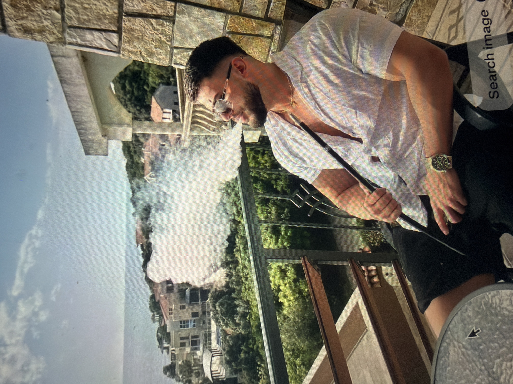 The image displays a male individual, appearing to be in his late 20s or early 30s, seated on an outdoor balcony. He has short dark hair, a trimmed beard, and wears reflective rectangular sunglasses. His attire consists of a loose-fitting, short-sleeved white or light-colored collared shirt, unbuttoned at the top to reveal a gold-colored chain with a small, flat pendant around his neck. A prominent gold-toned wristwatch with a dark face is visible on his left wrist. He is actively exhaling a significant volume of white vapor or smoke from a black hookah hose, which he holds to his mouth with his right hand. He is seated in a dark chair, with his left leg resting near a grey, textured table that displays a computer mouse cursor, indicating the image is a photograph of a digital screen.
The balcony features a dark metal railing in the foreground and a textured stone-tiled pillar or wall to the right, consistent with exterior architecture. The background presents a panoramic view of a coastal landscape. Below the balcony, there is a dense expanse of lush green vegetation, interspersed with numerous multi-story residential buildings featuring various architectural styles and tiled roofs, extending towards the coastline. Beyond the immediate coastal development, a calm, vast body of water, identifiable as the sea or ocean, stretches to the distant horizon under a mostly clear, light blue sky with sparse white clouds. The geographical features depicted are characteristic of a coastal environment.