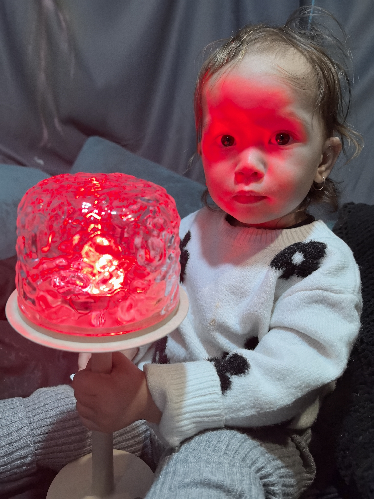 A young child is holding a glowing red, round, table lamp with a white base and stem. The child is seated, wearing a white sweater with black flower-like markings, and gray knit pants. The child's face is illuminated in red light from the lamp. The background appears to be an interior space with pillows and drapery. The exact location in the city of Noviy, Russia cannot be determined from the image.