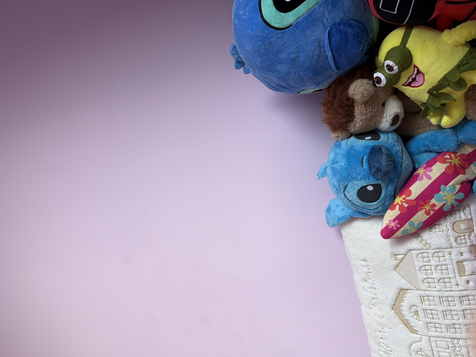 This is a close-up, eye-level, indoor, medium shot of several plush toys arranged against a pale purple wall. The plush toys include a large blue Stitch character, a smaller blue Stitch character holding a pink and yellow surfboard, a brown bear or lion character, a yellow Minion character with goggles and a wide smile, and a portion of a red plush toy that appears to be from the Squid Game. The toys are clustered together in the lower right portion of the frame. To the left of the toys is a white, fuzzy textile with a design of houses and the text "Merry Christmas" embroidered in gold thread. The lighting is soft and appears to be coming from the left, creating subtle shadows. The image does not contain any recognizable landmarks or features that would indicate its location in the city of Etropole, Bulgaria.