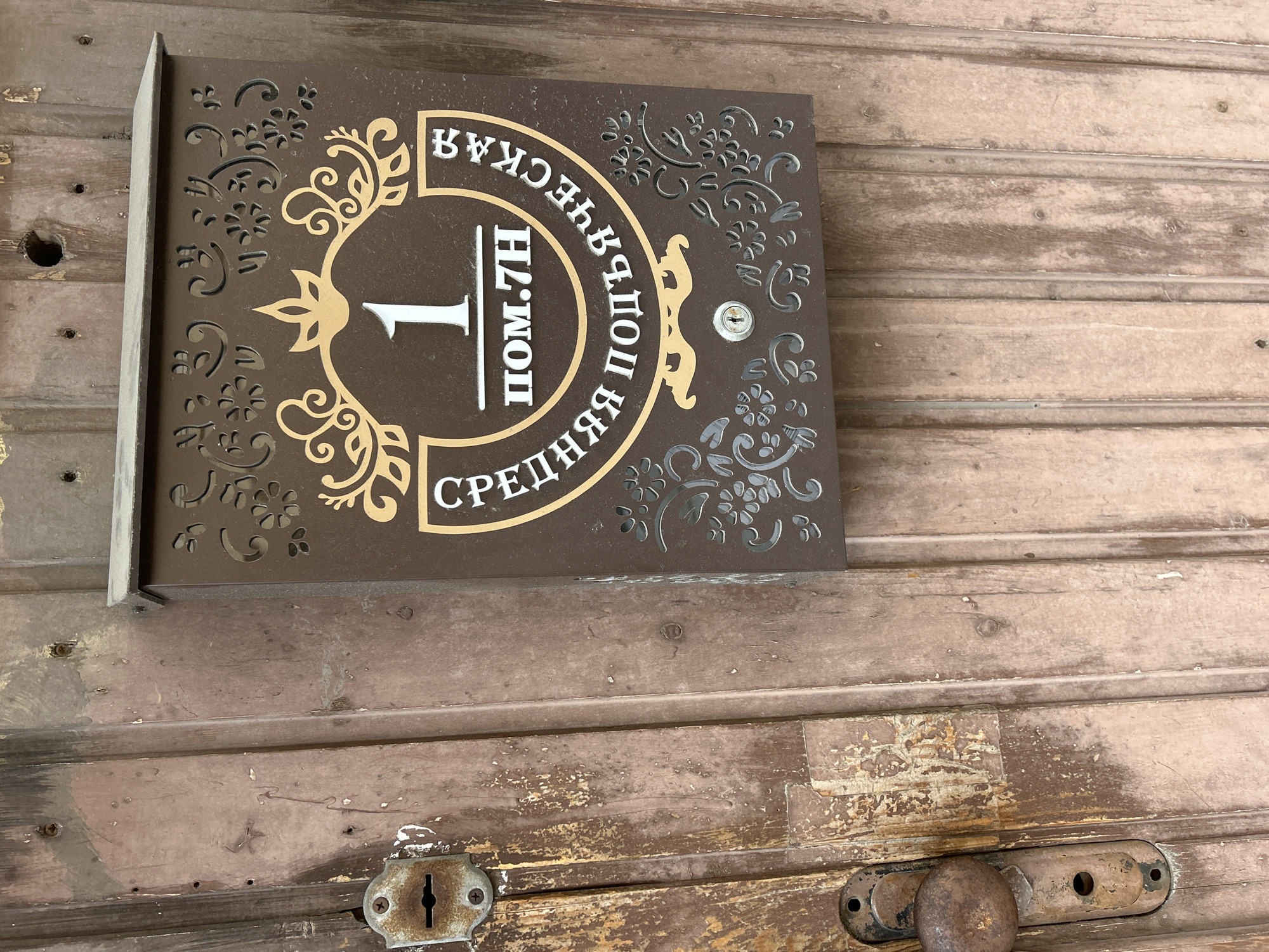 This media file shows a close-up, eye-level shot of a decorative sign mounted on a weathered wooden door. The sign is rectangular with rounded corners and is brown with ornate gold and white lettering and patterns.
The prominent subject is the sign itself, which appears to be an address or office marker. It features a large white number "1" in the center, surrounded by a gold, decorative oval. Above the "1," white Cyrillic text reads "ПОМ.7H," indicating apartment or office number 7H. Arched text in white Cyrillic runs around the top and sides of the oval. The top text, read counter-clockwise, says "ВНЕШНЯЯ" (Vneshnyaya), and the text on the right, also read counter-clockwise, says "СРЕДНЯЯ ПОЩ" (Srednyaya Poshch). The overall design suggests an older building.
The sign is attached to a wooden door that shows significant signs of age and wear. The wood is rough-hewn and has a distressed, weathered appearance with peeling paint and visible grain. There are several horizontal planks making up the door. Below the sign, near the bottom right of the frame, are two metal door hardware elements: a rusty keyhole and a portion of a door handle mechanism, both appearing old and heavily used.
The lighting suggests daytime, with no strong shadows indicating a specific time of day. The scene is likely outdoors, given the wooden door and hardware, but the immediate surroundings are not visible. The overall impression is one of age, history, and perhaps a bit of disrepair, yet the sign itself retains a decorative charm.
