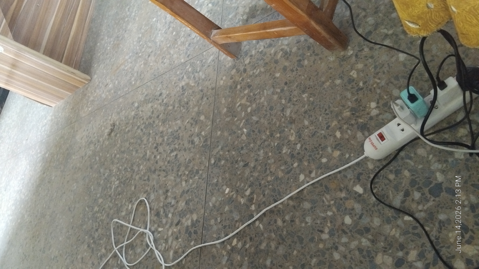 A white power strip with multiple plugs, including a light blue adapter, is lying on a speckled gray floor. A white cord extends from the power strip upwards and to the left, looping loosely. A black cord also connects to the power strip and extends downwards and to the right. Part of a wooden chair or table leg is visible on the right side of the image. The floor appears to be indoors, possibly in a residential or commercial space in Howayi, Nigeria. The timestamp "June 14, 2026 2:13 PM" is present in the bottom left corner. There are no people visible in the image.