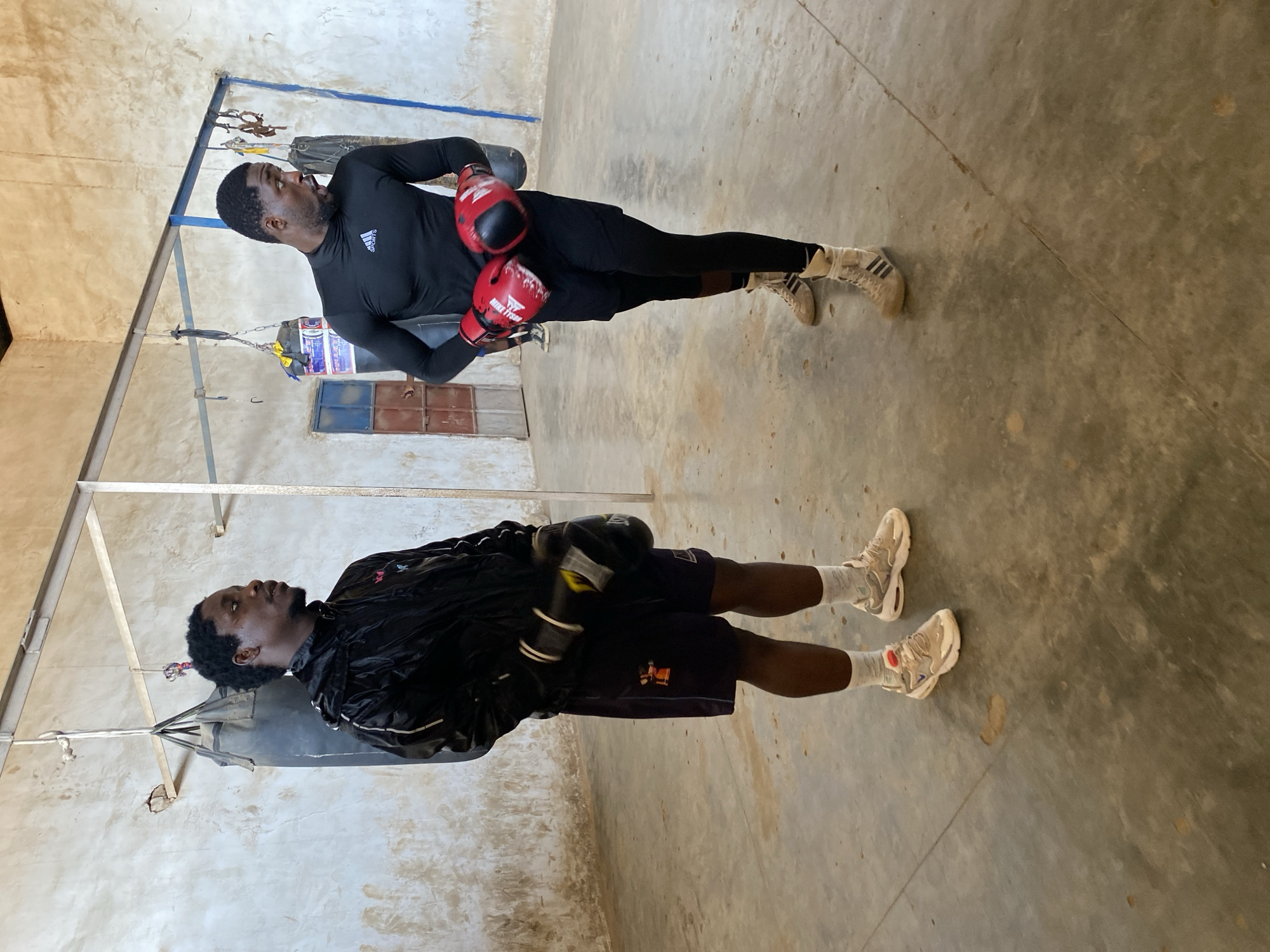 Two young black African men are visible in an indoor training area, possibly a gym, located in Jos, Nigeria.
One man, wearing a black long-sleeved Adidas shirt and black leggings, is positioned at the top of the image. He has red boxing gloves on his hands and tan-colored Adidas sneakers on his feet.
The second man is located below him, near a hanging heavy punching bag. He is dressed in a shiny black jacket, dark shorts, and white socks, and he has light-colored sneakers on his feet.
The walls and floor of the space appear to be concrete. There is a metal frame visible in the background. Natural light is present. A small window with blue frames is visible in the background.