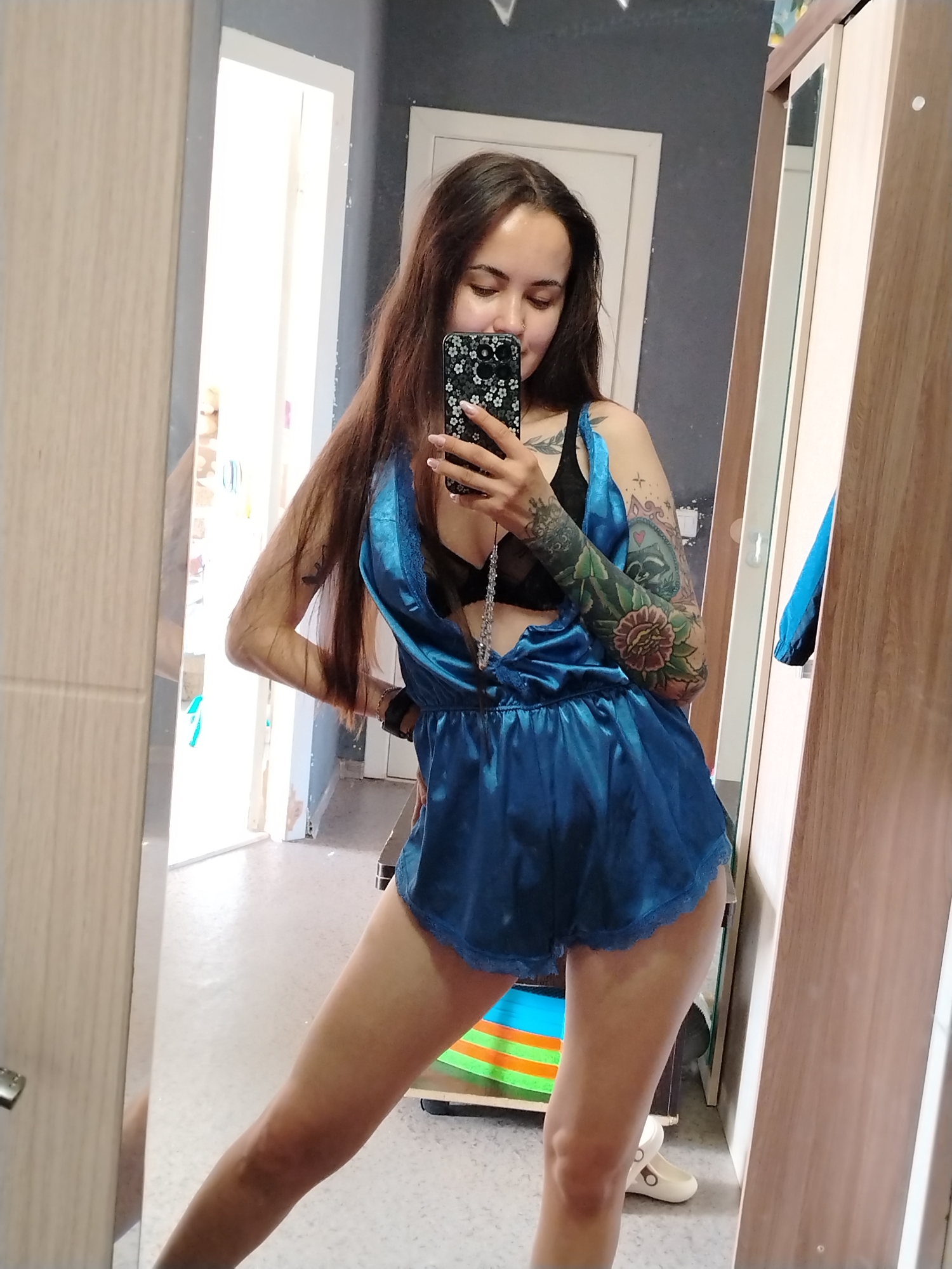 A woman is taking a selfie in a mirror. She has long brown hair, a black bra, and a blue satin romper. She has extensive tattoos on her left arm. The room appears to be a bedroom with a mirror, wardrobe, and doorway. The city of Noviy, Russia is not discernible from the image.