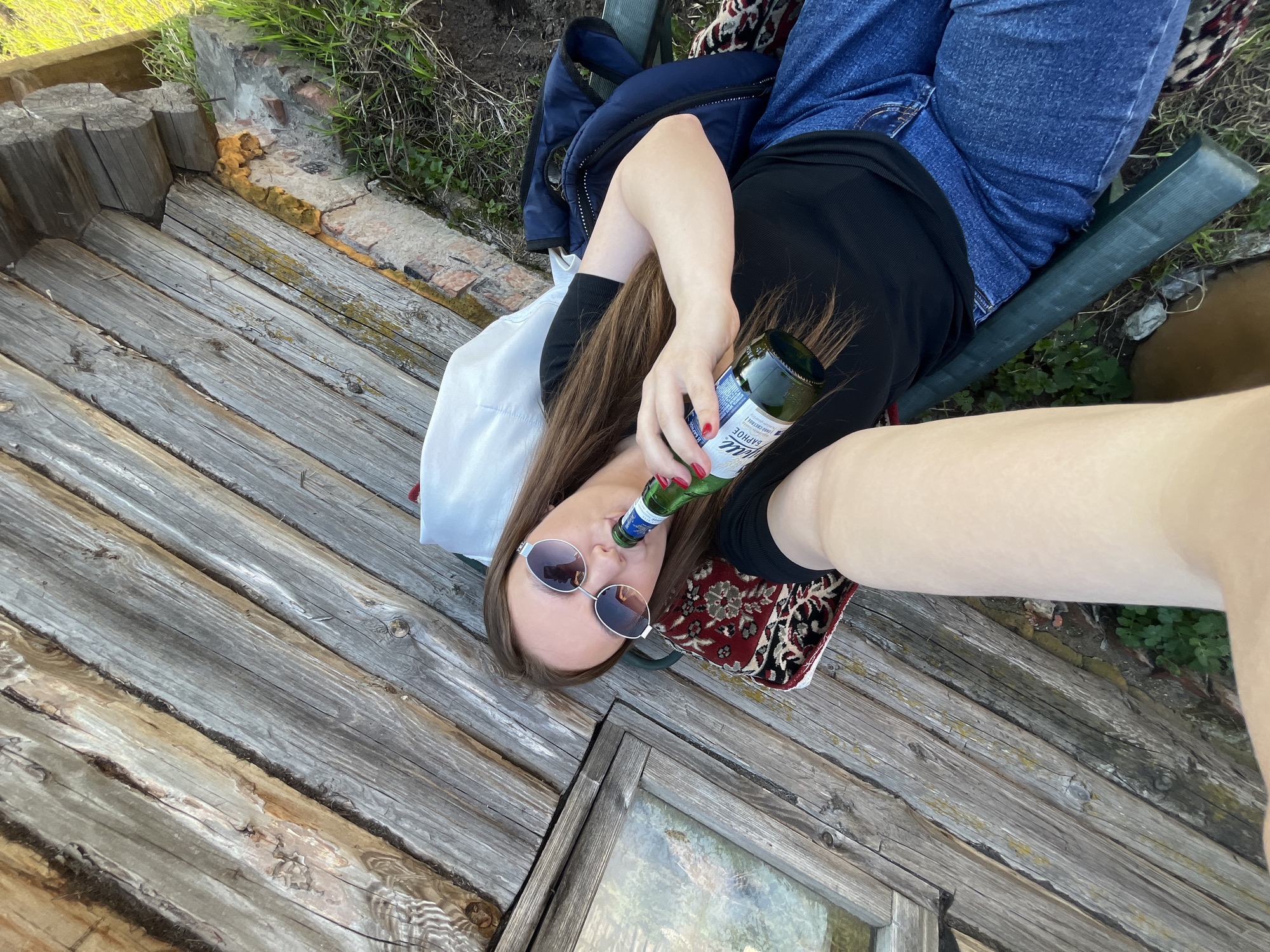 A woman lies on a weathered wooden surface, possibly a deck or platform. She is wearing sunglasses and holding a green bottle to her lips, as if drinking. She has on a black top, blue jeans, and her nails are painted red. A dark jacket lies behind her. Her left arm extends toward the camera, nearly filling the right side of the image. The location is likely Chaikovsky, Russia, based on the Cyrillic label on the bottle, a green-bottled beer sold in that country.