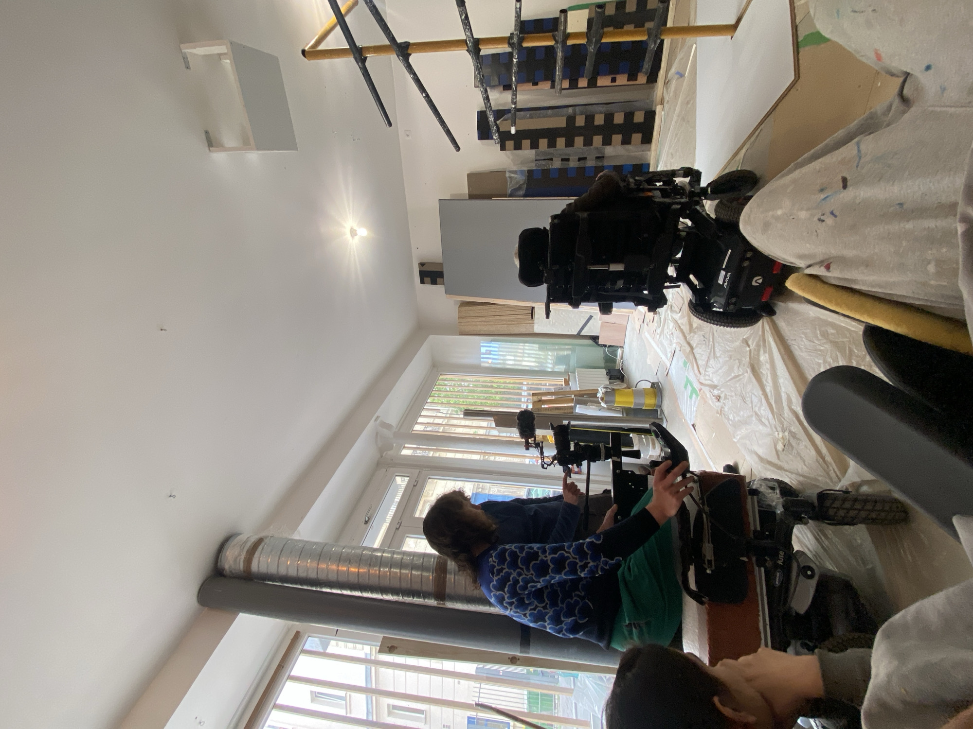 This media file captures an indoor scene in what appears to be a large studio or workshop, possibly located in Paris, France. The room is brightly lit by natural light streaming through expansive windows that span one wall, revealing views of other urban buildings outside. The floor is covered with protective plastic sheeting, and numerous boards and canvases, some painted with geometric patterns in blue, black, and brown, are visible in the background, suggesting an ongoing art or set design project.
The image features at least three individuals and two prominent powered wheelchairs. One person, with shoulder-length brown hair and wearing a blue and white patterned top with green trousers, is seated in a wheelchair facing the windows. Their hands are engaged with a control device connected to a camera or recording equipment mounted on a specialized rig. Another person is partially visible behind them, also seemingly focused on the equipment. A third individual is seen in the foreground on the right, wearing a grey hoodie splattered with paint, and appears to be involved in the activity or preparation.
Visible objects include two high-tech powered wheelchairs, one with "AVIVA" and another with "Kite" branded on its side. The camera setup includes an external microphone. In the background, there are various stacked wooden boards, a yellow paint roller, and a yellow canister. A roll of silver corrugated tubing and a grey pipe run along the wall near the windows. Above, a structure of wooden and metal bars, possibly scaffolding or a lighting rig, is present. A single bare lightbulb hangs from the ceiling, and a small white rectangular structure is mounted on the wall/ceiling.
The overall scene suggests a creative or production environment, likely involving a film shoot or a recording session within an art studio, with preparations for painting or construction also evident. The individuals appear focused on their tasks. The presence of the specialized wheelchairs indicates adapted equipment is in use for the activity.