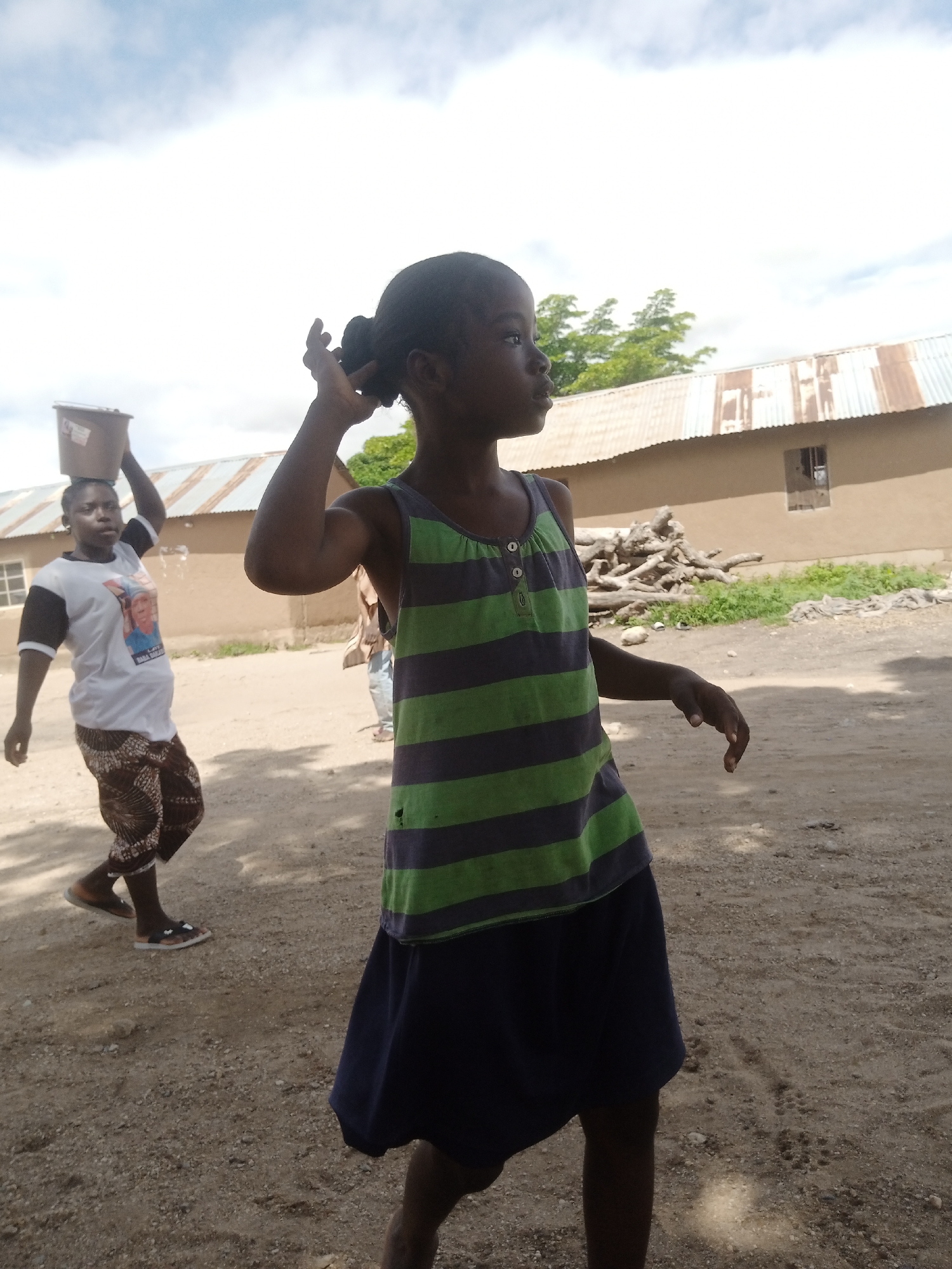 A young Black girl in a green and purple striped tank top and a navy blue skirt stands in a dusty outdoor area, her right arm raised as if adjusting her hair. She is positioned in the foreground, facing right and looking away from the camera. Behind her, a Black woman wearing a white t-shirt with an image on it and patterned pants walks from left to right, carrying a brown bucket on her head. In the background, a tan building with a corrugated metal roof and a small window is visible, with a pile of logs and green vegetation near its base. The ground is sandy and uneven, with scattered pebbles. The sky above is mostly white with some clouds. The scene appears to be in a rural or semi-urban area, possibly in Langtang, Nigeria, given the architectural style of the building and the general setting.