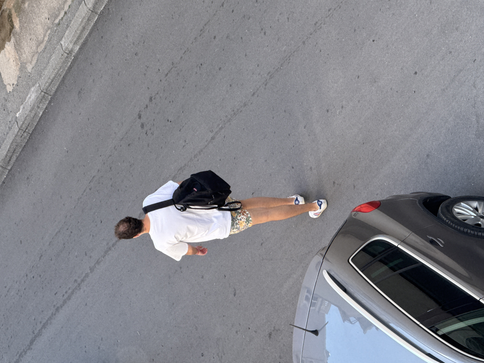 A top-down view shows a man standing on an asphalt road, next to a gray car. The man has short, dark hair and is wearing a white t-shirt, patterned shorts, and white sneakers with red and blue accents. He carries a black backpack. The gray car occupies the right side of the image, with a visible spare tire on the roof. The asphalt road takes up most of the remaining space. The image was likely taken in a city setting, given the presence of the road, car, and what appears to be a sidewalk along the top edge of the frame. Based on general architectural knowledge and the appearance of the asphalt road, the location could be Budva, Montenegro, or a similar European city, due to the visible architectural elements.