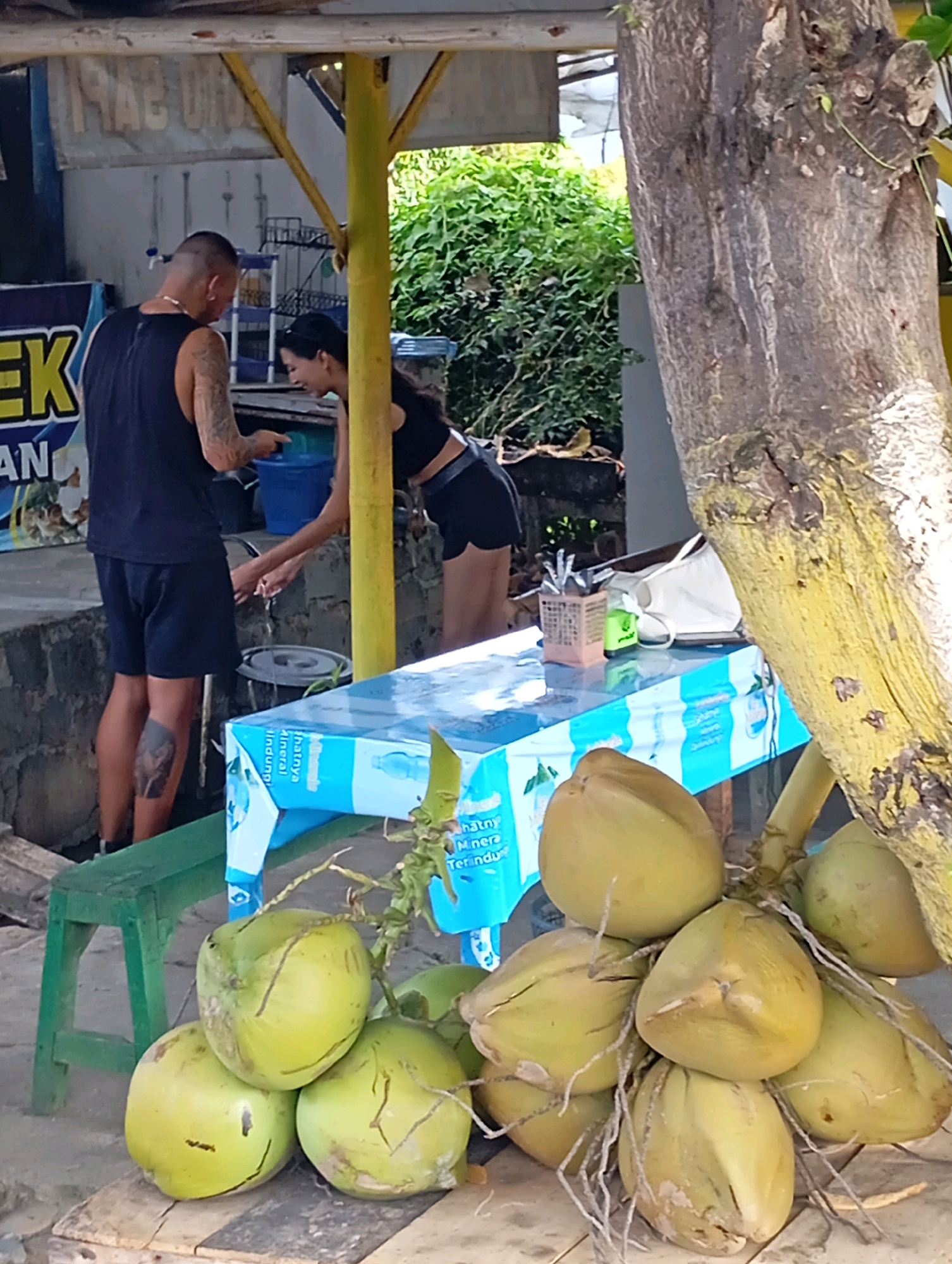 The image depicts a scene outside a food stall in Wanurejo, Indonesia. In the foreground, a cluster of green coconuts is displayed on a wooden surface. A man wearing a black tank top and shorts, with a tattoo on his left calf, stands to the left, facing a woman. The woman, dressed in a black crop top and shorts, is engaged in washing her hands under a faucet. To the right of the scene, a large tree trunk with textured bark is prominent. A blue and white plastic-covered table is situated between the people and the coconuts, holding a basket of utensils and a white bag. In the background, the stall structure is visible with yellow supports, and lush green foliage can be seen.
