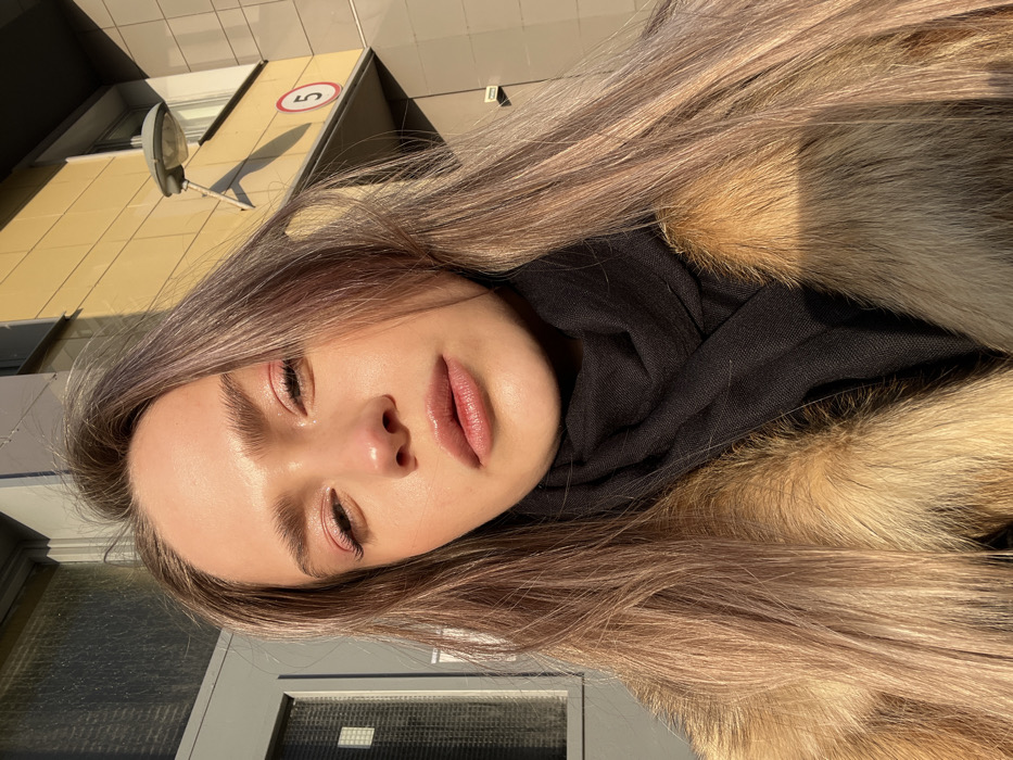 The image presents a close-up, chest-up portrait of a female subject standing outdoors in Saint Petersburg, Russia. The subject, facing the camera directly, exhibits long, light brown hair with a subtle purple-grey tint, styled down. Her face features prominent eyebrows, visible pink eyeshadow, and full, glossy lips slightly parted. She is dressed in a black scarf wrapped around her neck, layered beneath a light brown or reddish-brown fur coat. Strong sunlight illuminates her face from the viewer's left, creating highlights and casting shadows. The background consists of a modern building facade: a dark grey section with a reflective, multi-paned window or door is visible to the subject's left, while a light yellow or beige paneled section extends to her right. On the yellow facade, a grey, wall-mounted street light fixture with a curved arm is present, positioned above a white circular sign with a red border containing the black numeral "5". Shadows from the light fixture are distinctly visible on the yellow wall, indicating direct sunlight.