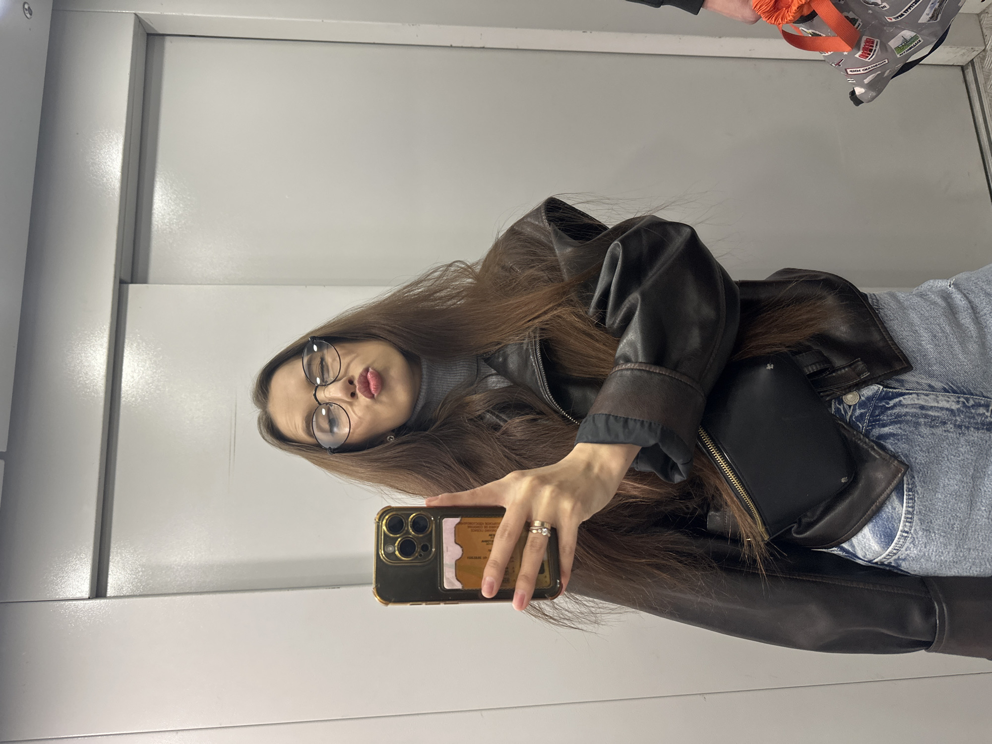 A young woman is taking a selfie in an elevator. She is wearing a brown leather jacket, gray turtleneck, blue jeans, and eyeglasses. She has long brown hair. She is holding a gold-colored smartphone. The background is the interior of an elevator with white walls. An orange bag with a black and white patterned design is visible in the top right corner. It is impossible to ascertain that the location is Yekaterinburg, Russia from the image.