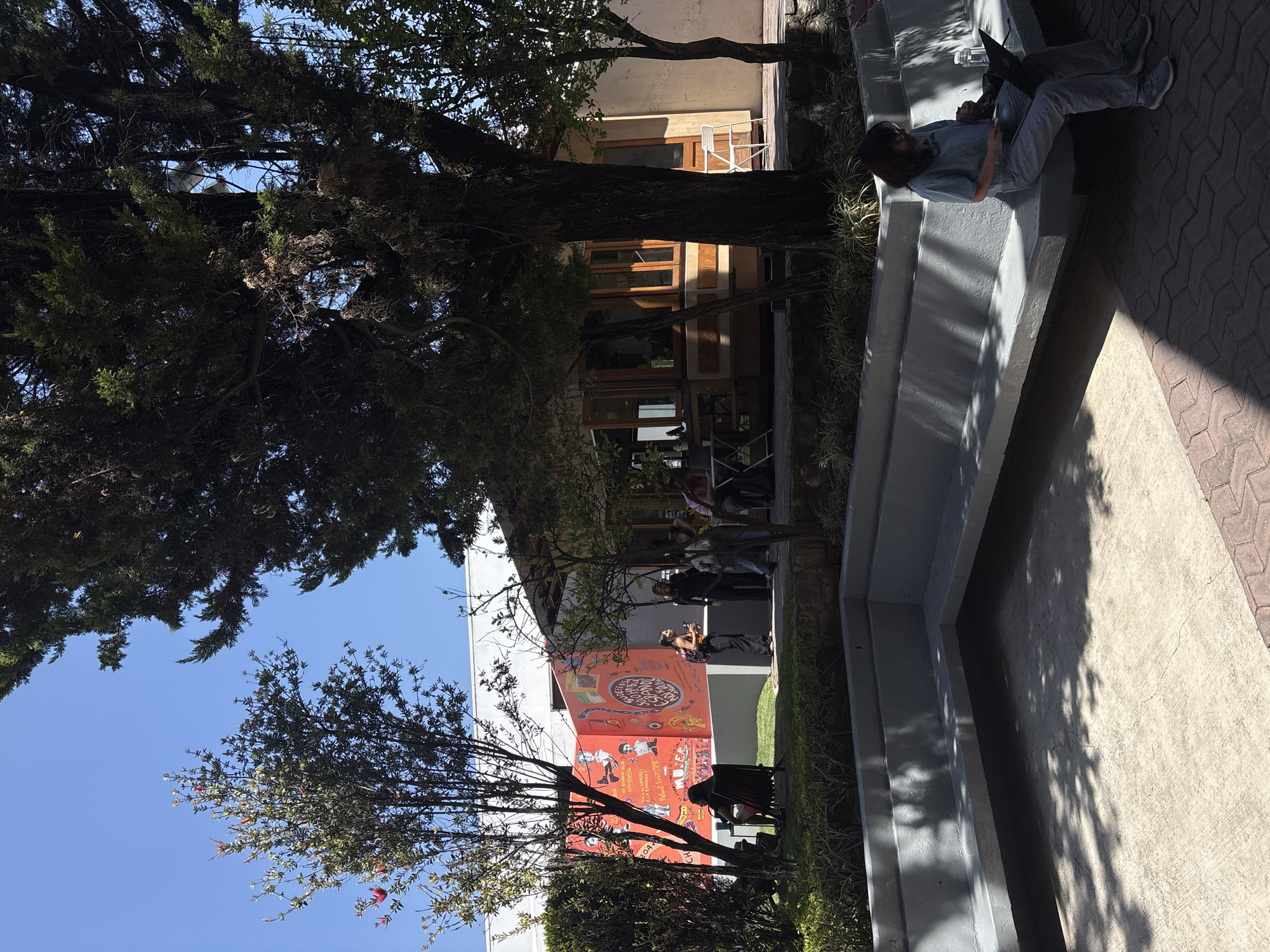 The image depicts an outdoor scene in San Andrés Cholula, Mexico, possibly on a university campus or a similar public space. A young person is sitting on a concrete structure that appears to be a bench or a low wall, using a laptop computer. The person is dressed casually in light-colored clothing.
Behind the bench, there are trees, and further back, a building with large windows and a colorful mural. The mural is predominantly red and features illustrations and text in Spanish. Additional people are present in the background near the mural, some of whom appear to be taking photos. The scene is illuminated by sunlight, casting shadows on the ground. The ground is a mix of concrete and brick paving.