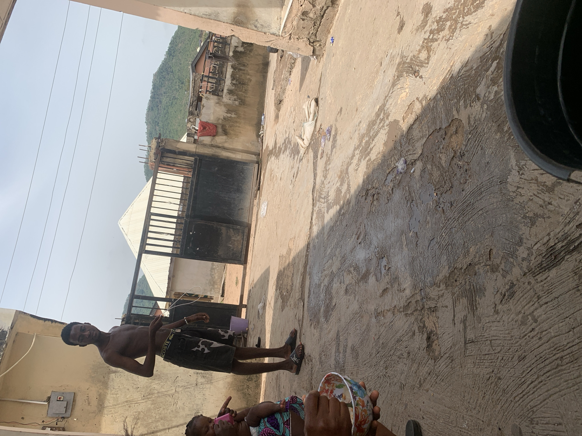 The image depicts a scene in Lokoja, Nigeria. A shirtless young man wearing shorts and sandals stands on a weathered concrete surface, seemingly in a courtyard or outdoor area. A small child lies nearby, being attended to by an individual holding a colorful bowl or cup. In the background, there's a metal gate, buildings, and a distant, vegetated hill. A black container is visible in the upper right corner. The concrete surface appears worn and stained.