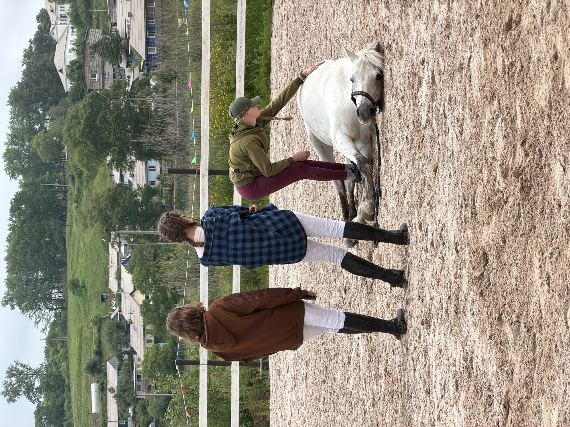 In Теплоозёрск, Russia, on a sandy equestrian ground, a white horse lies on its side. Two young women are present, one standing to the left, facing away from the camera, wearing a brown hooded jacket and white pants. The other woman, also facing away, is in the center, dressed in a blue plaid shirt and white pants. A third individual, wearing a green hooded jacket and maroon pants, is kneeling beside the horse, gently touching its back. The background features a white fence, a field of yellow flowers, and a hillside dotted with houses and trees, indicative of a rural or semi-rural setting. The sky is overcast.