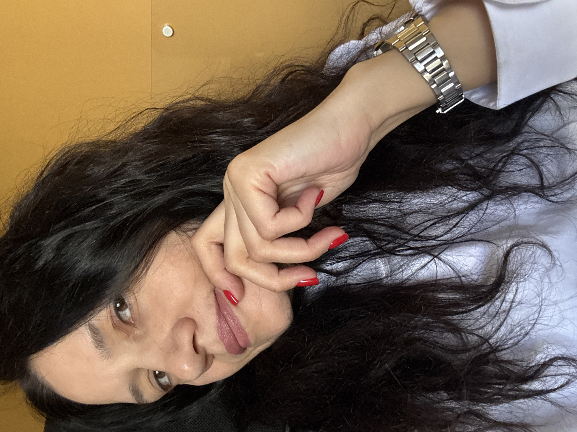 A close-up, tilted shot shows a woman with long, dark, wavy hair. Her face is partially obscured by her hand, where her fingernails are painted red. She wears a silver metal watch on her wrist, and a white garment is visible. The background features a light-yellow wall with a small, white circular object affixed to it. There is no indication of the city of Chaikovsky, Russia in the image.