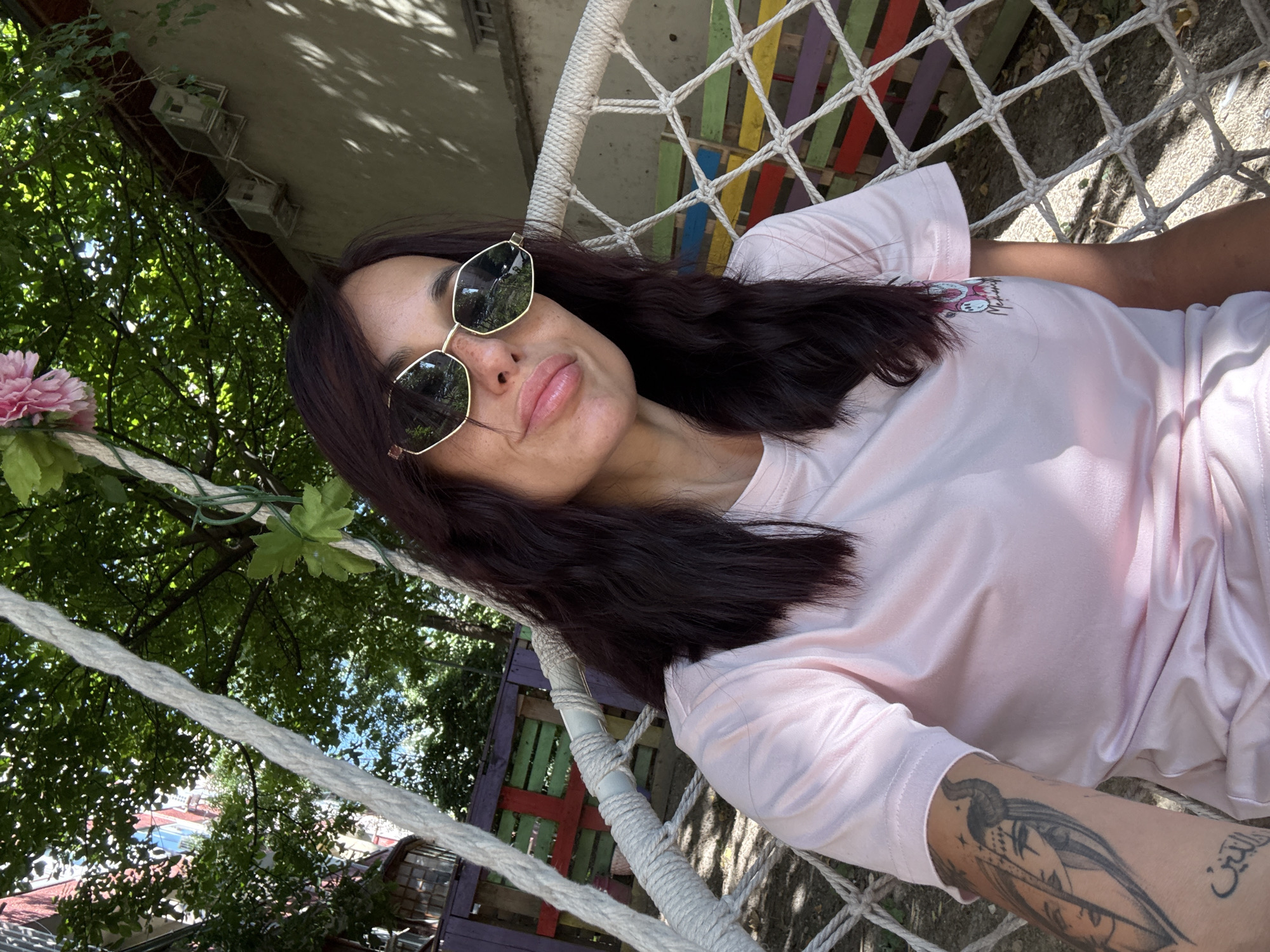 A woman is taking a selfie while sitting on a hammock swing. She has dark hair and is wearing sunglasses and a pink t-shirt with a cartoon graphic. She has tattoos on her arm. The hammock is hanging from a tree branch, and a colorful wooden fence is visible in the background. The scene appears to be outdoors in a residential area, with buildings and trees surrounding the hammock. The overall impression is a casual, relaxed setting on a sunny day. The location is the backyard of a house in Pleven, Bulgaria.