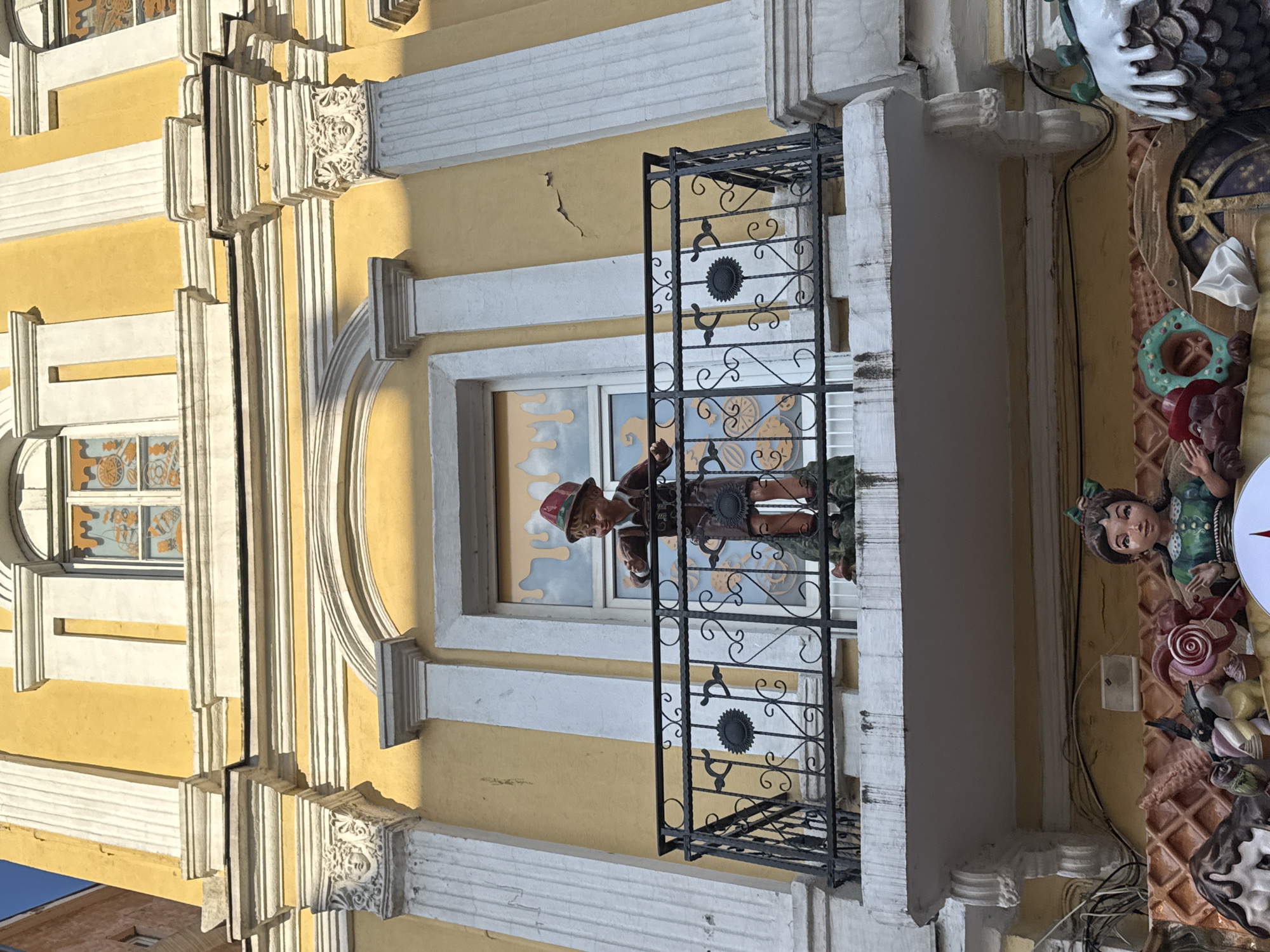 The image captures a whimsical scene on the exterior of a building, likely in Plovdiv, Bulgaria. The building has a yellow façade with white architectural detailing, including columns, window frames, and decorative moldings. A small balcony with an ornate black metal railing juts out from the building.
On the balcony stands a life-sized figure of a boy wearing a red hat and traditional attire. He appears to be holding a cup. Behind him, a window displays a whimsical, cartoon-like painting of clouds and other figures.
To the right of the balcony, the building's façade is adorned with a variety of confectionery-themed decorations, including a human figure, lollipops, waffles, and other sweet treats. Wires are running along the building façade.