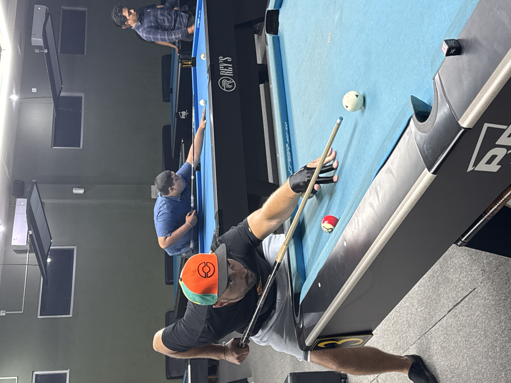 An indoor billiards hall in Kuta Selatan, Indonesia, features at least three individuals engaged in playing or observing a game of pool. In the foreground, a Caucasian male wearing a black t-shirt, grey shorts, a distinctive orange and green baseball cap, and a black glove on his left hand is poised to strike a white cue ball with a pool cue. His focus is intensely directed towards the ball. Adjacent to the cue ball is a red and white striped pool ball. Another player, a person of Asian descent, is visible in the middle ground, wearing a blue t-shirt and leaning over a pool table. In the background, a third individual, wearing a plaid shirt, stands near another pool table. The environment is characterized by blue pool tables, overhead lighting fixtures, and dark green walls. The overall impression is one of a recreational activity taking place in a dedicated sporting venue.