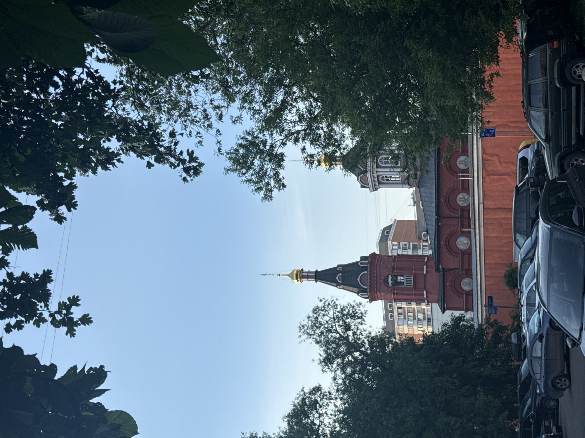 The image shows a building with a red facade and a dark spire topped with a golden dome, partially framed by trees. The building appears to be a church or religious structure, possibly located in Moscow, Russia. The image is taken from a low angle, looking up at the building. The building's design and coloration suggest it is a historical building, possibly in the Byzantine or Russian Revival style. There are cars parked along the right side of the image. Modern apartment buildings are visible behind the church. The sky is a pale blue.