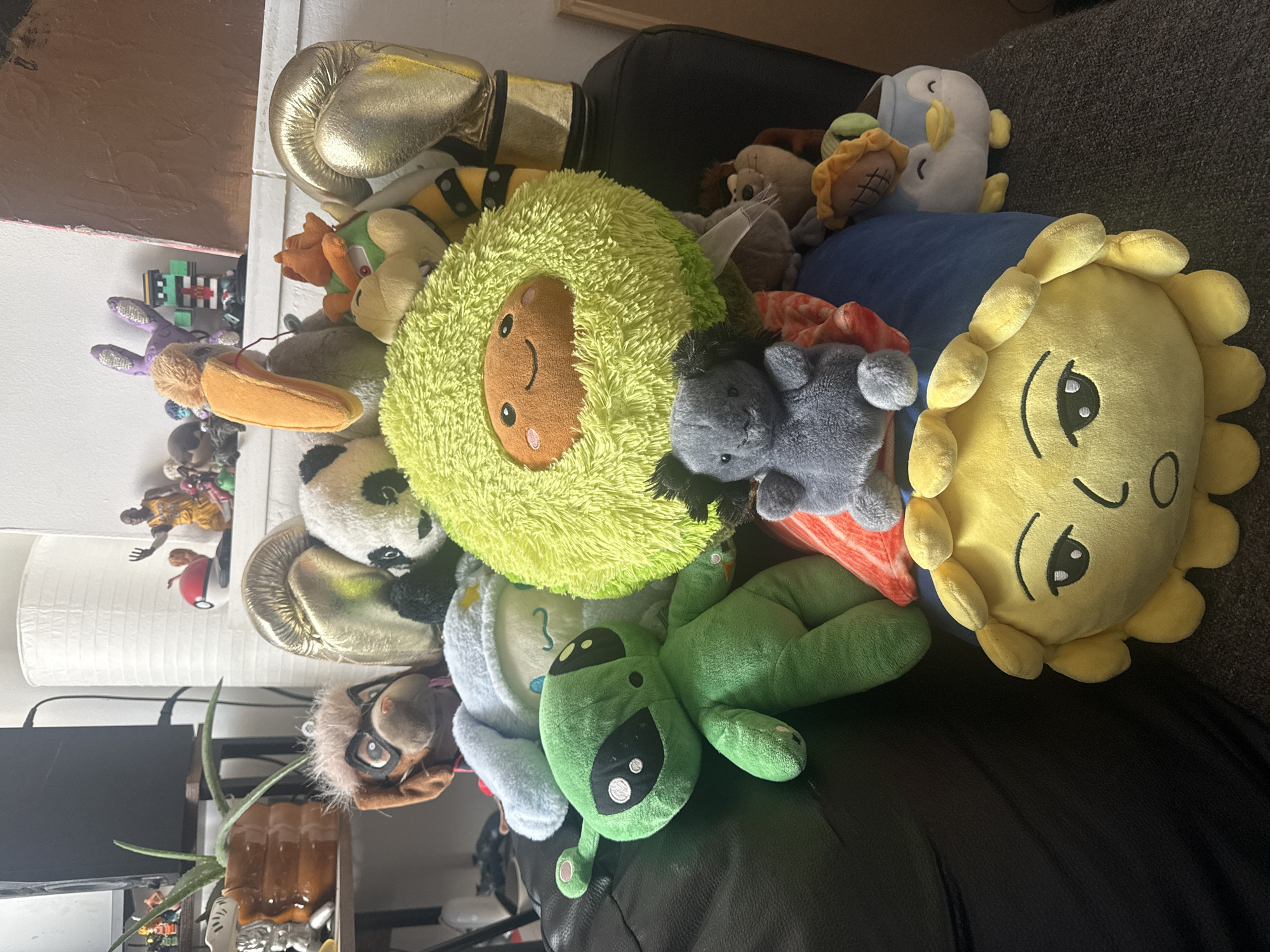 The image shows a pile of plush toys on a dark surface, likely a couch or chair. The plush toys include an avocado, a green alien, a grey donkey, a yellow sun, a penguin, and a panda. There are also several other toys that are difficult to identify due to being partially obscured by the other toys. A gold boxing glove is near the top of the pile. The environment shows a shelf and a wall. The location cannot be determined from the image.
