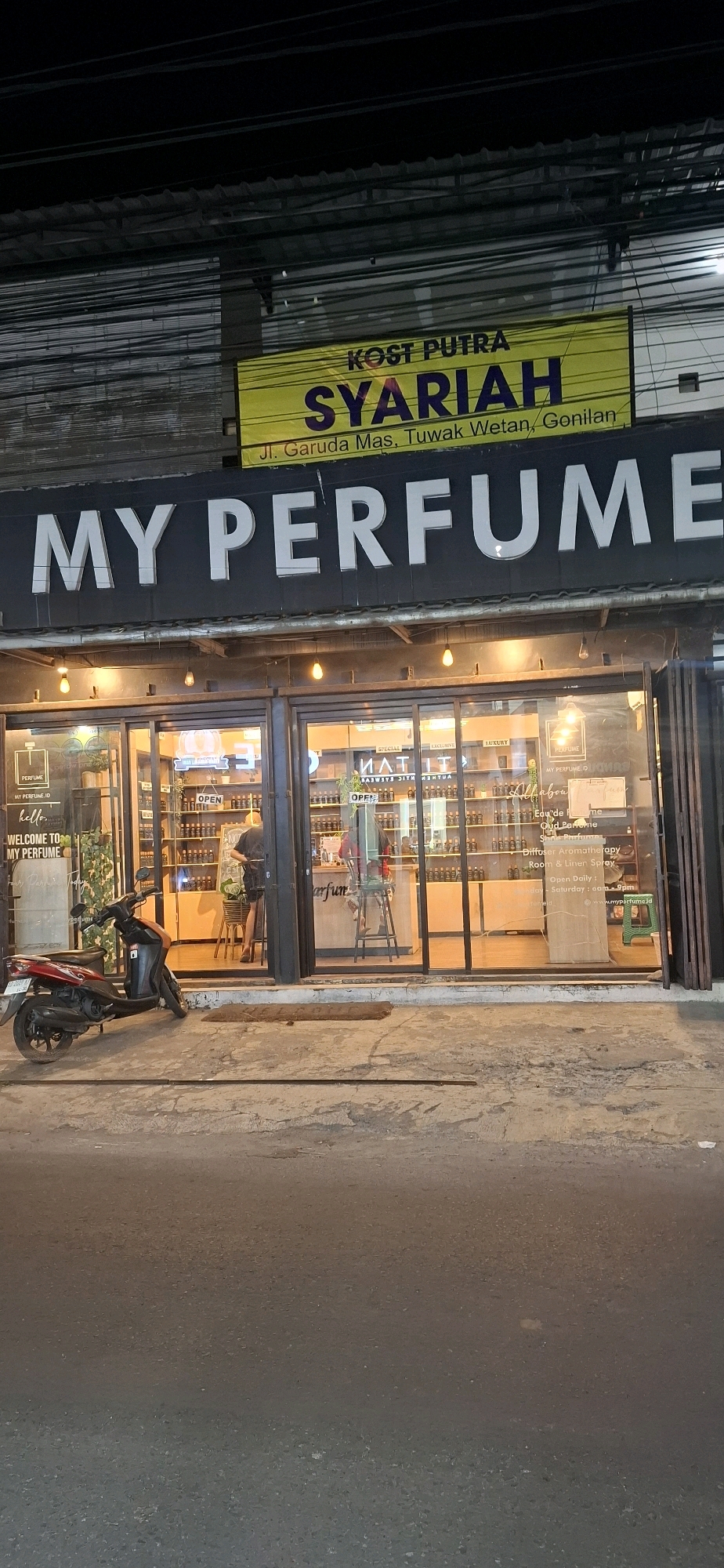 This is a nighttime photograph of a street-level commercial establishment in Pabelan, Indonesia. The primary subject is the storefront of "MY PERFUME", a business selling perfumes. The store features large glass windows displaying rows of perfume bottles on shelves. Above the windows, a prominent sign in white sans-serif capital letters reads "MY PERFUME". A yellow banner above the "MY PERFUME" sign displays text in Indonesian, including "KOST PUTRA SYARIAH" and an address: "Jl. Garuda Mas, Tuwak Wetan, Genilan".
In front of the store, a red and black scooter is parked on the pavement. The entrance to the store is open, and an individual is visible inside, appearing to be working behind a counter. The street in front of the store is paved and appears wet, suggesting recent rain or damp conditions. The lighting is artificial, with warm yellow lights illuminating the interior of the store and some exterior lighting visible. Overhead, a complex network of electrical wires is visible against the dark night sky. The scene is likely from a commercial or mixed-use area of Pabelan, Indonesia.