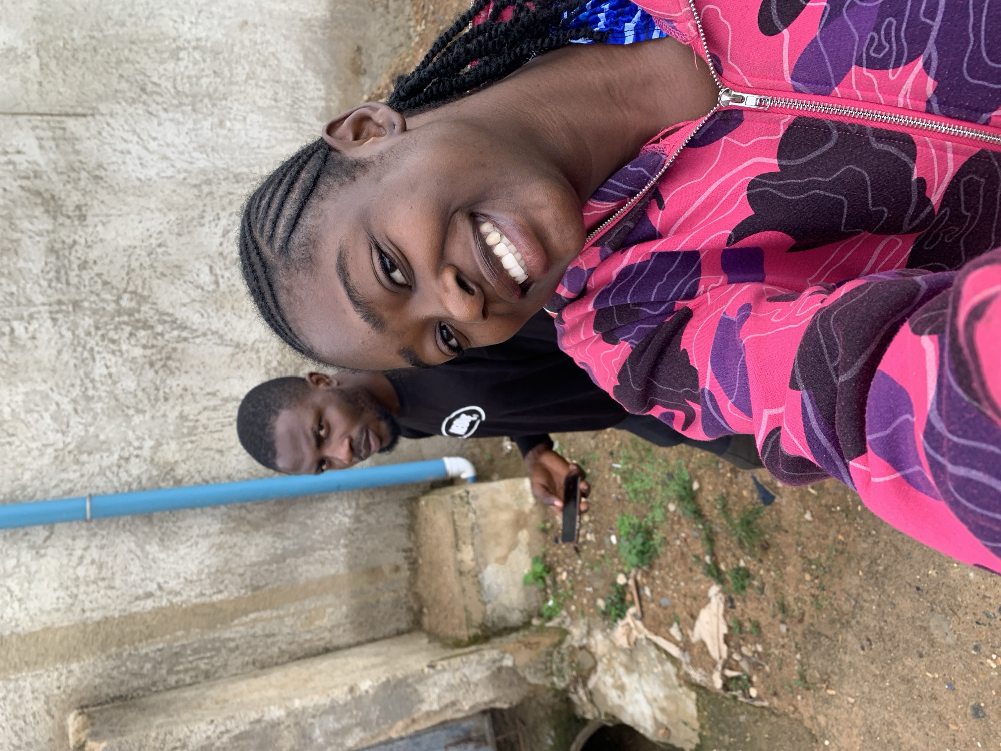 The image depicts two individuals in an outdoor setting in Rafin Par, Nigeria. In the immediate foreground, a dark-complexioned female is captured in a selfie-style shot, smiling broadly and exposing white teeth. Her hair is styled in cornrows at the front leading into braids. She is wearing a vibrant pink hooded jacket with a purple and dark grey camouflage-like pattern, featuring a metallic zipper running down the front. A blue and white patterned fabric is visible beneath her jacket collar. Behind her, to the left, stands a dark-complexioned male with a beard, looking towards the camera with a serious expression. He is wearing a black long-sleeved shirt with a white circular logo on the left chest and is holding a black smartphone in his right hand.
The background features a light grey, textured concrete wall with vertical dark streaks indicating wear. A prominent vertical light blue pipe is affixed to this wall by a metal strap, descending to a white elbow joint connected to a lower pipe section. The ground is uneven, composed of sandy brown earth, small stones, and sparse green vegetation. Several rough concrete blocks or foundations are distributed across the ground, and an open, dark concrete conduit or drain is visible on the far left side of the scene. Light-colored debris is scattered on the ground.