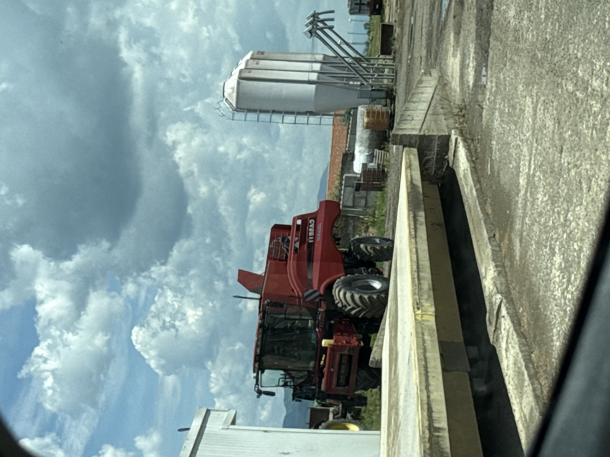 The image depicts agricultural equipment and structures, potentially a farm scene. A large red combine harvester is visible, along with a cylindrical grain silo supported by metal scaffolding. The background shows a cloudy sky. The scene appears to be next to a concrete structure. No people are visible. The precise location is specified as Straldzha, Bulgaria.
