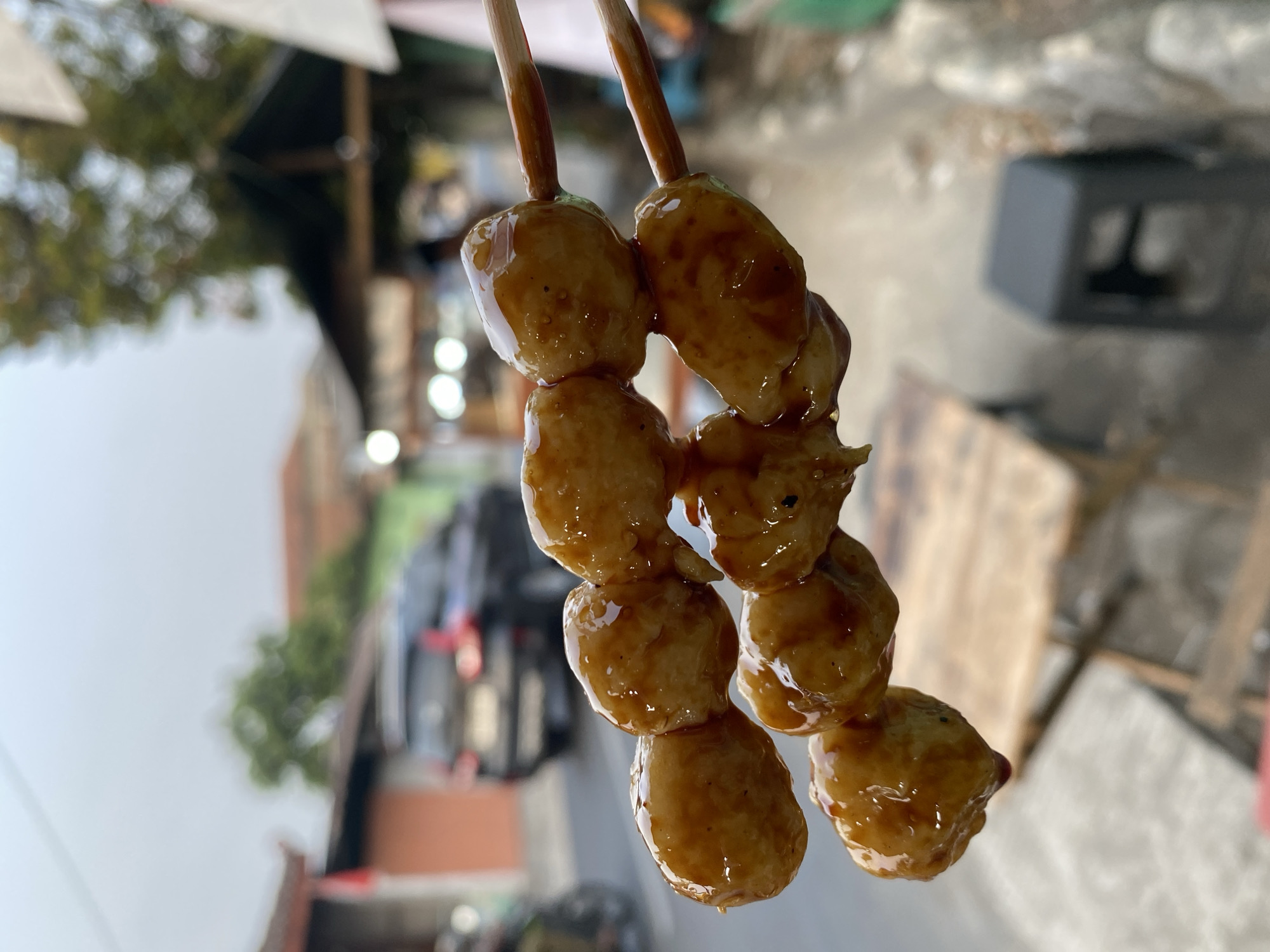 A close-up, eye-level shot focuses on two skewers of glistening, golden-brown meatballs, coated in a dark, glossy sauce. The meatballs are arranged in two staggered rows, with four visible on the top skewer and three on the bottom. They appear to be cooked and sauced, with some sauce dripping from the bottom skewer. The skewers are held horizontally by two wooden chopsticks, positioned in the center of the frame.
The background is blurred, indicating a shallow depth of field, and suggests an outdoor setting, likely a street food stall in Kasihan, Indonesia. Visible elements in the background include a dark-colored SUV, a red-tiled roof on a building, a dark-colored canopy, and other indistinct structures that suggest a market or roadside environment. The sky is overcast, giving the scene a soft, diffused light. A small, dark plastic stool is visible in the lower right corner of the frame. The overall impression is a casual, perhaps humid, day in a bustling urban or semi-urban area.