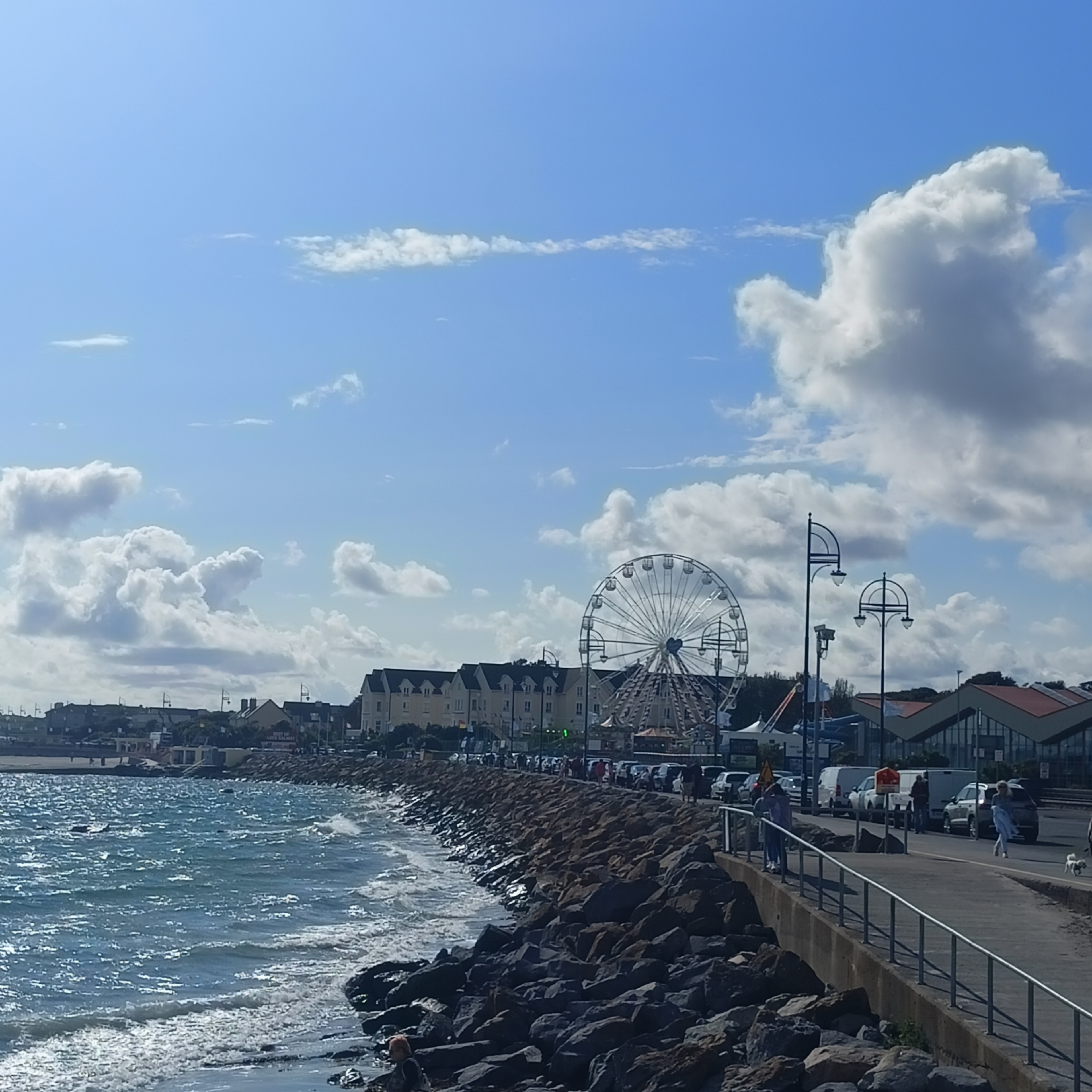 The image captures a bright, sunny day at the Salthill Promenade in Galway, Ireland. The foreground is dominated by the choppy, blue waters of Galway Bay, with waves gently lapping against a rocky shoreline. A concrete promenade runs along the coast, bordered by a metal railing, and is populated by several individuals. A woman walks a small white dog, while another person, possibly a child, is further ahead. Cars are parked along the promenade. In the middle ground, a large Ferris wheel stands prominently, suggesting a funfair or amusement area. Behind the Ferris wheel, a row of cream-colored buildings with dark roofs lines the promenade. Further in the distance, more buildings and structures are visible. The sky is a vibrant blue, dotted with fluffy white clouds, indicating pleasant weather. The scene conveys a lively atmosphere typical of a seaside resort on a clear day.