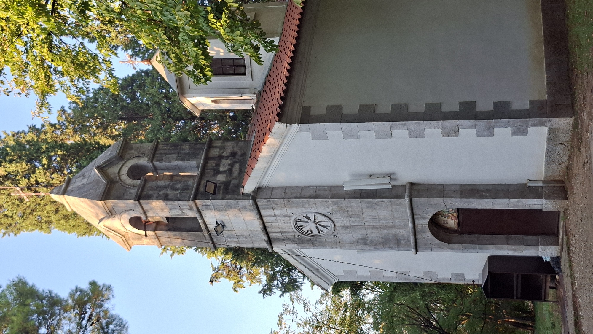 A stone bell tower and a white church with a red-tiled roof are visible. The bell tower has arched openings and a bell inside. A stone wheel-like carving is present on the tower. The church has a dark brown door framed by an arched stone entrance. There is a small dark structure to the left of the entrance. Lush green trees surround the church. The image appears to be taken in the daytime. This is the Church of Saint John the Baptist in Podgorica, Montenegro.