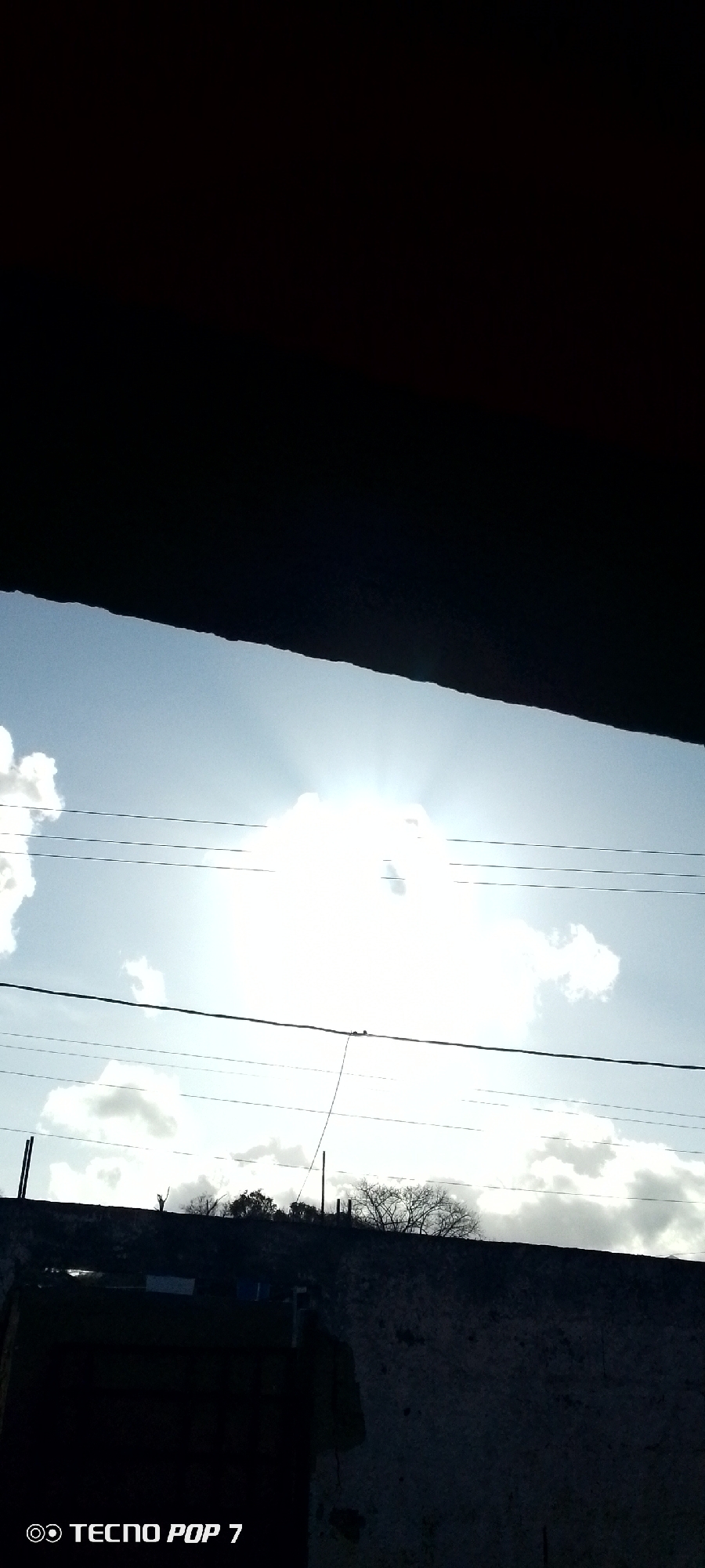 The image shows a view of the sky with clouds and the sun obscured by one large cloud, casting rays of light. Overhead power lines are visible across the frame. The foreground is partially obscured by a dark overhanging structure, and a wall with some vegetation and trees on top of it can be seen. The photo was taken with a Tecno Pop 7 phone. No people are visible in the image. There is no information in the image to confirm it was taken in Altagracia, Venezuela.
