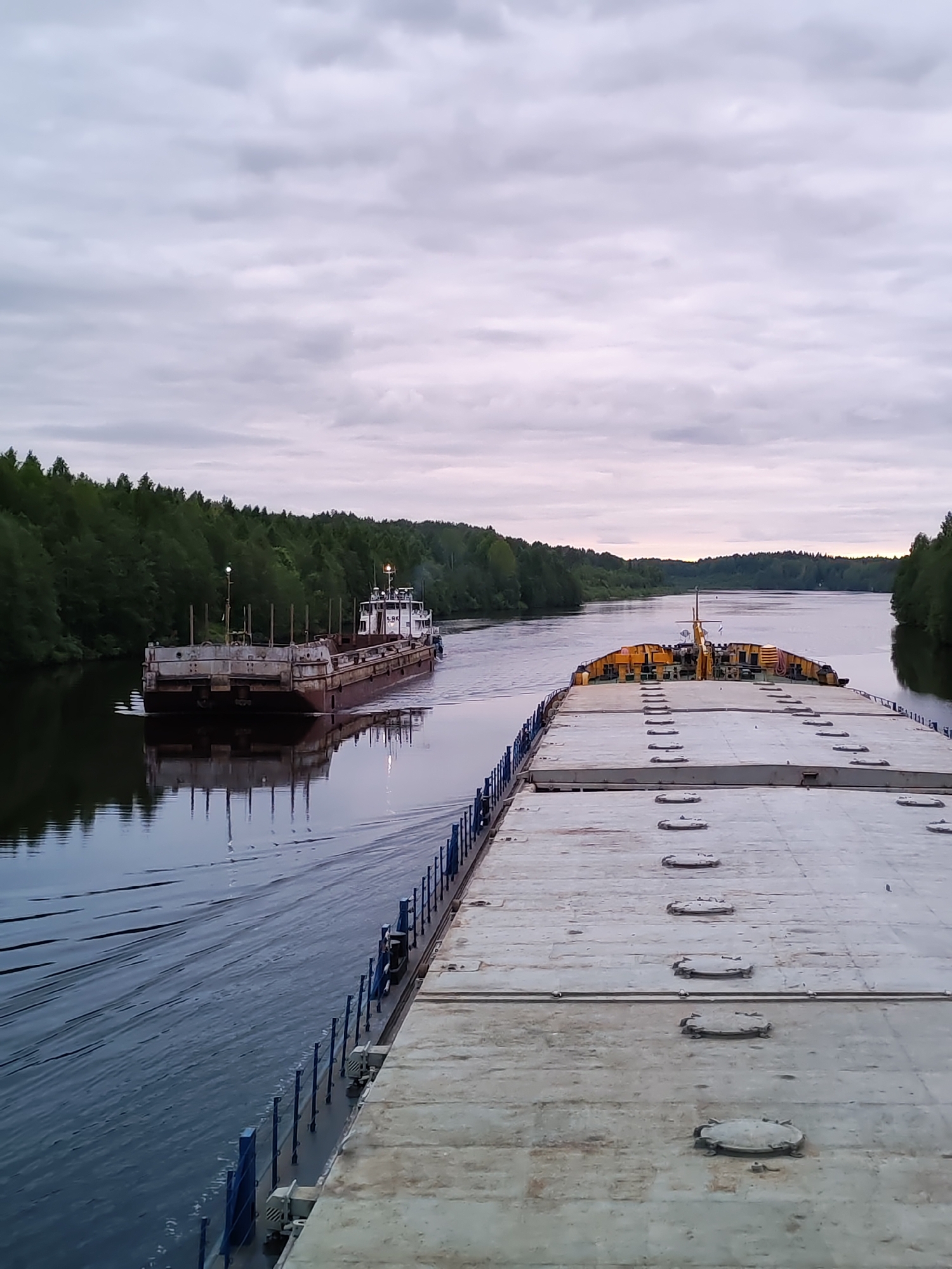 The image shows a river scene in Volokov Most, Russia, featuring two vessels navigating a waterway. The foreground is dominated by a large, flat-topped barge with a grey concrete surface and regularly spaced hatches. A blue railing lines the edge of the barge. In the midground, another vessel, possibly a tugboat or smaller barge, is visible. It has a brown hull, a white superstructure, and several vertical posts along its deck. The water is dark and reflects the vessels and the surrounding trees. The river is bordered on both sides by dense, green forest. The sky is overcast with muted colors, suggesting early morning or late evening light.