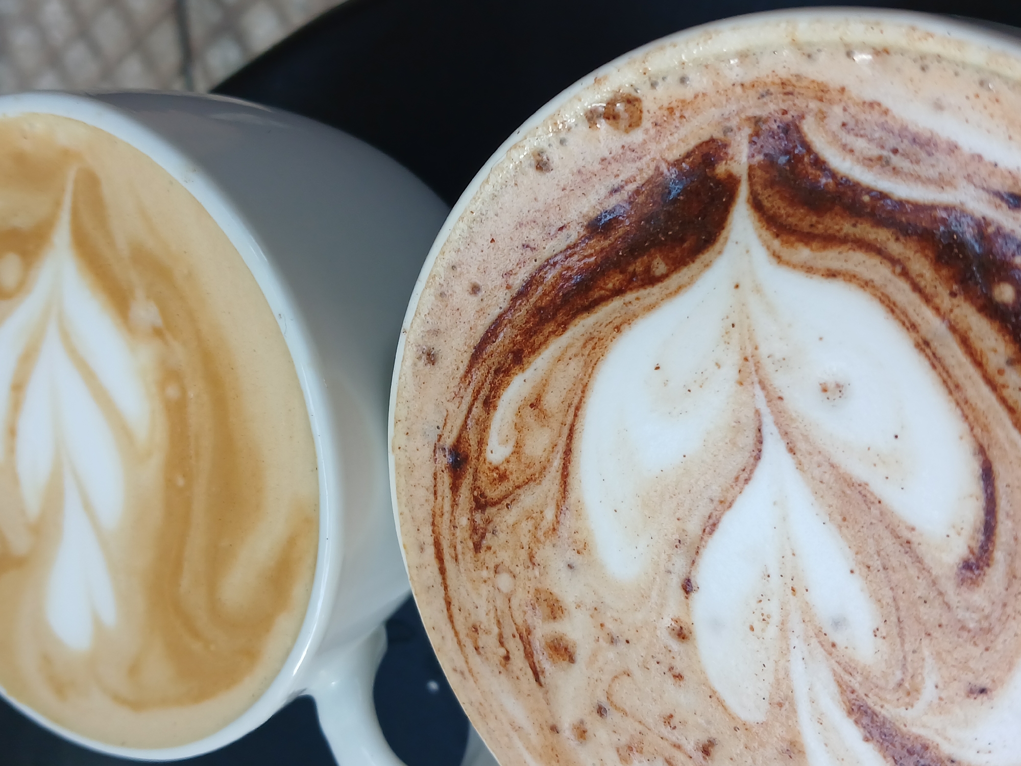 The image shows two white ceramic mugs filled with latte-style coffee drinks on a dark, possibly black, surface. Both drinks have latte art. The mug on the right has a darker, more mocha-colored foam, with a distinct dark brown pattern creating a stylized floral or heart shape. The mug on the left contains coffee with a lighter brown hue and a similar, though less defined, latte art pattern. The setting appears to be indoors, likely a cafe or restaurant. There are no people visible. The image does not contain information to determine its location in Buenos Aires, Argentina.