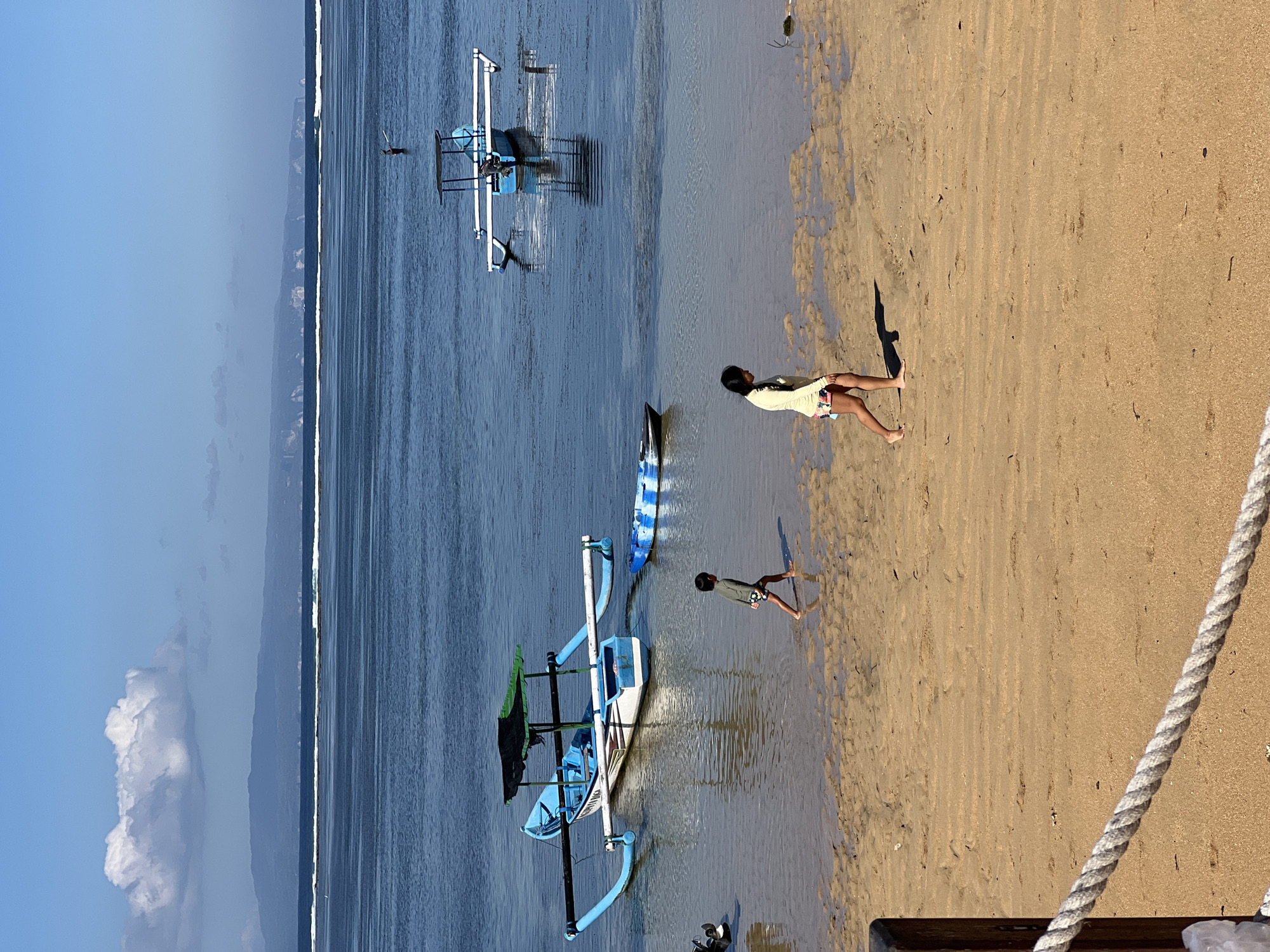 The image captures a serene beach scene in Denpasar, Indonesia. Two children are present, one a young girl in a light-colored top and patterned shorts, and a younger boy in a green shirt and shorts. The girl is standing near the water's edge, her hands clasped in front of her, while the boy walks along the sandy shore.
Several traditional outrigger boats are visible in the shallow water. One is a prominent blue and white vessel, while another, with green netting, rests closer to the shore. A surfboard with a blue and white pattern lies next to the second boat. The beach is composed of light brown sand, marked with footprints and ripples from the receding tide.
The water is a calm, deep blue, reflecting the clear sky. In the distance, a hazy coastline with mountains can be seen, suggesting a picturesque tropical setting. The sunlight casts distinct shadows on the sand, indicating it is likely late afternoon. The overall atmosphere is peaceful and relaxed, evoking a sense of a typical day at a Indonesian beach. There is no visible text in the image.