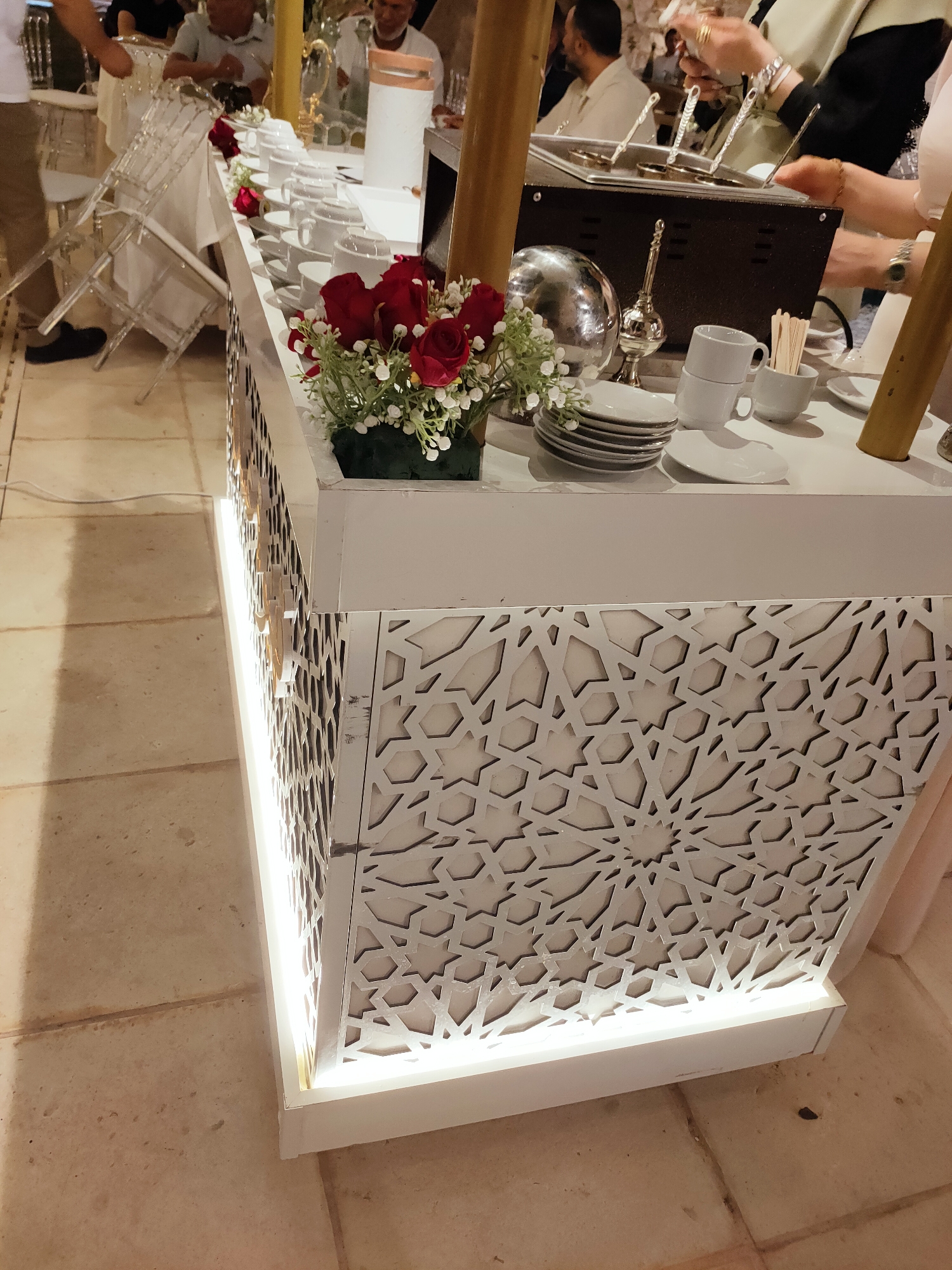 The image depicts a buffet or serving station set up for an event, likely a reception or meal, in Cebalat, Tunisia. The station is decorated with intricate white geometric patterns on its facade and illuminated by LED lights along its base. On the table, there are stacks of white plates, several white cups and saucers, and a bouquet of red roses with white filler flowers. Behind the plates and cups, a hot beverage dispenser with ladles is visible. In the background, several people are blurred, suggesting a gathering or event in progress. The flooring appears to be tiled with large, light-colored stones.