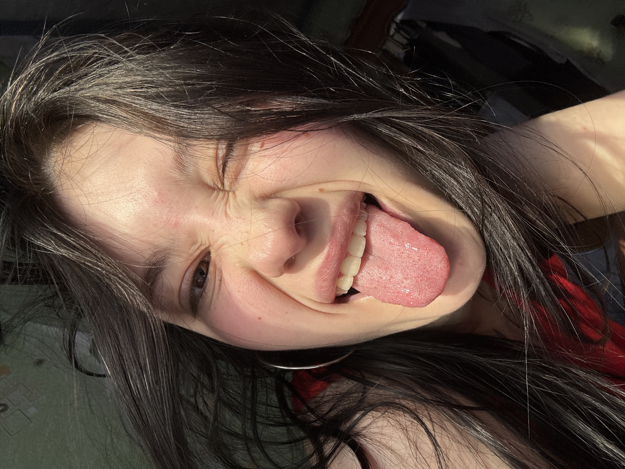 An extreme close-up depicts a young woman in what appears to be an indoor setting. Her face is centered, angled slightly to the right, and illuminated by strong, natural light from the right side. She has long, dark brown hair, part of which is visible around her face and shoulders. Her facial expression is a combination of a wink with her left eye (viewer's right), which is tightly closed and scrunched, and her mouth open with her tongue extended. The right eye (viewer's left) is open, showing a dark iris. A silver-colored hoop earring is visible on her left ear. Minor skin imperfections, including a few small red marks and faint freckles, are present on her face. A glimpse of red fabric is visible on her left shoulder area. The background is dark and indistinct, offering no identifiable features of a specific location or the city of Фоки, Russia.