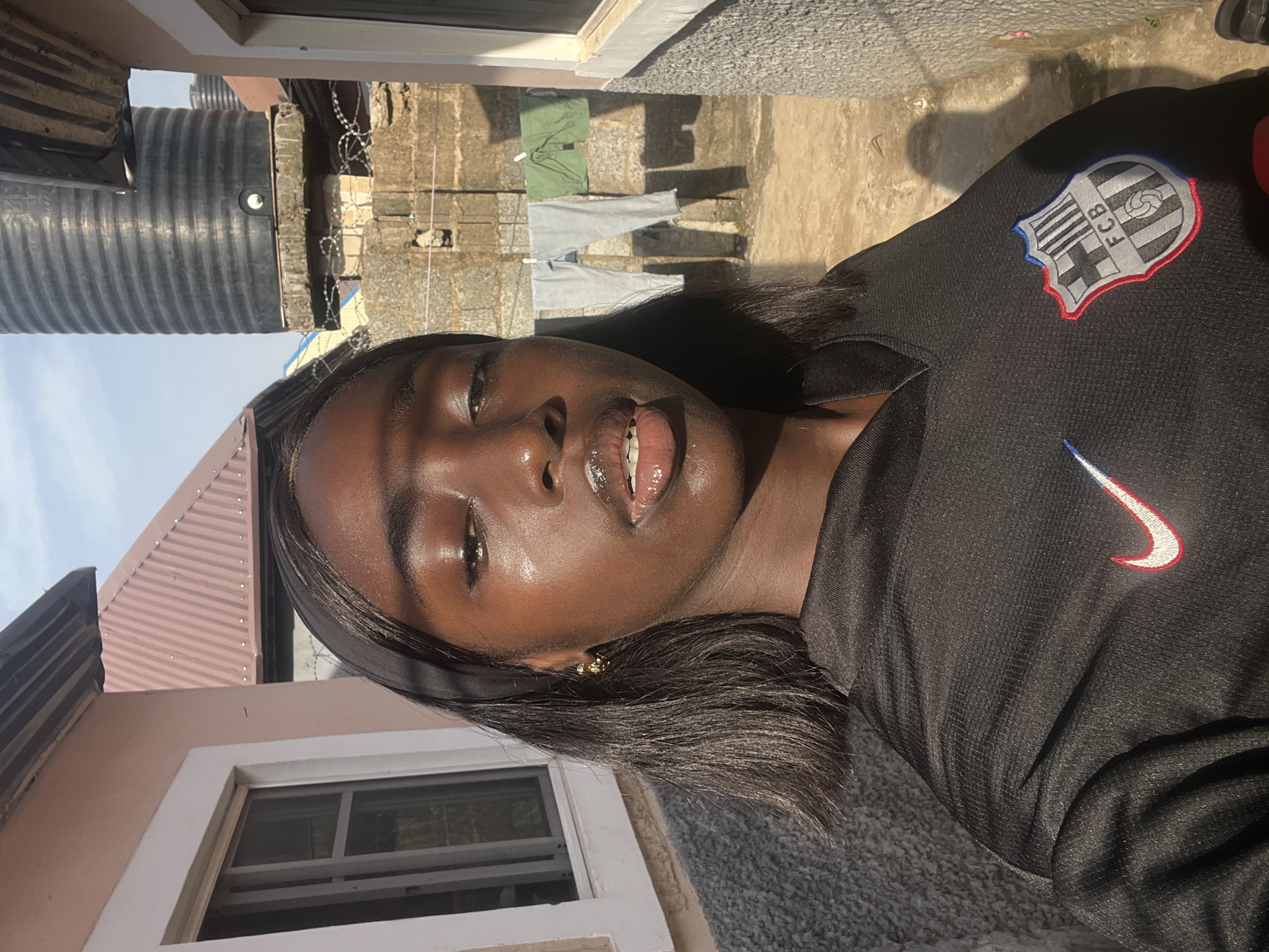 A young woman with dark skin stands outdoors. She is wearing a black shirt with a Barcelona FC badge on the upper left and a white Nike swoosh on the right. The background shows a building with a corrugated iron roof and a water tank. Clothes are hanging on a clothesline between buildings. The location appears to be in a residential area of Abuja, Nigeria.