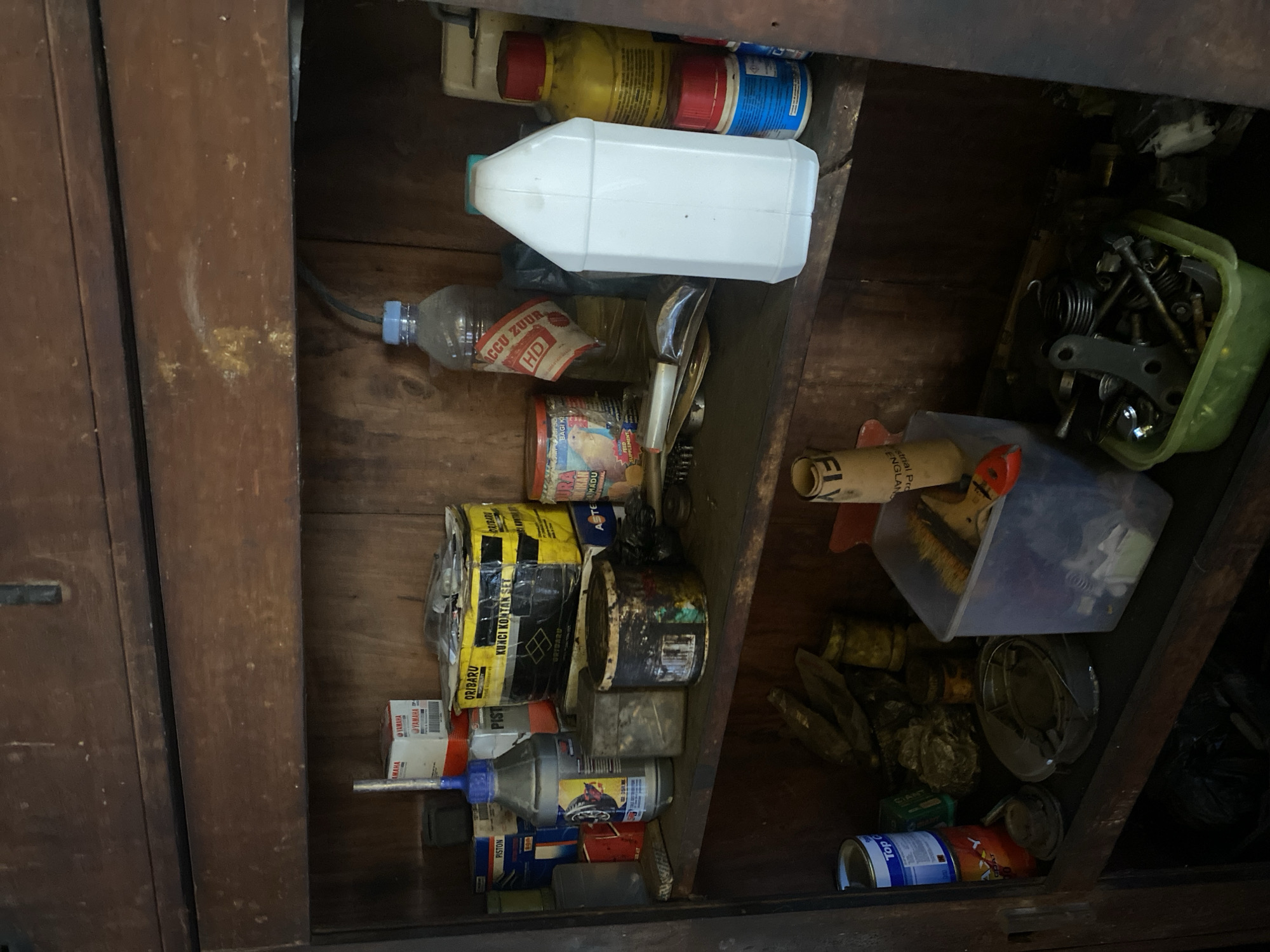 The image shows a wooden cabinet filled with various items, likely in a workshop or storage area. On the top shelf, there are several boxes, including some labeled "YAMAHA" and one large yellow box with "KUNCI KONTAK SET" written on it. There's also a bottle of liquid with a red label. The middle shelf contains more bottles, cans, and some metal tools. Notably, there is a large white plastic jug and a yellow bottle with a red cap. The bottom shelf is less organized, with cans, a plastic container filled with nuts and bolts, and other miscellaneous items. The overall environment appears to be cluttered and functional. The scene is located in Bantaeng, Indonesia.