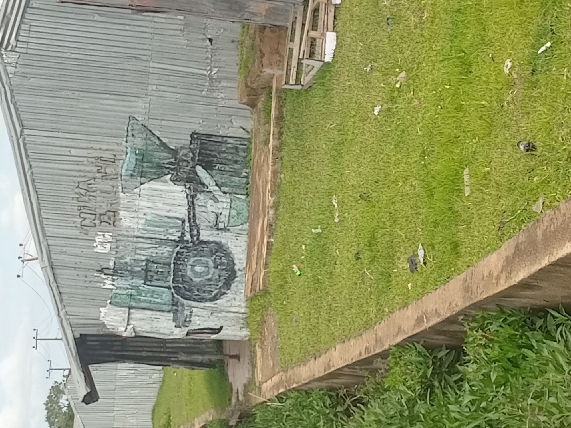 The image shows a corrugated metal building with graffiti depicting a farm machine. The building appears to be in a state of disrepair. In the foreground, there is a grassy area with some scattered debris, including plastic bags and paper. A concrete border runs along the left side of the image, separating the grass from a patch of green vegetation. In the background, power lines and poles are visible against a cloudy sky. The overall scene suggests an urban or semi-urban environment in Bukuru, Nigeria, characterized by informal structures and visible signs of economic activity or daily life. There are no people visible in the image.