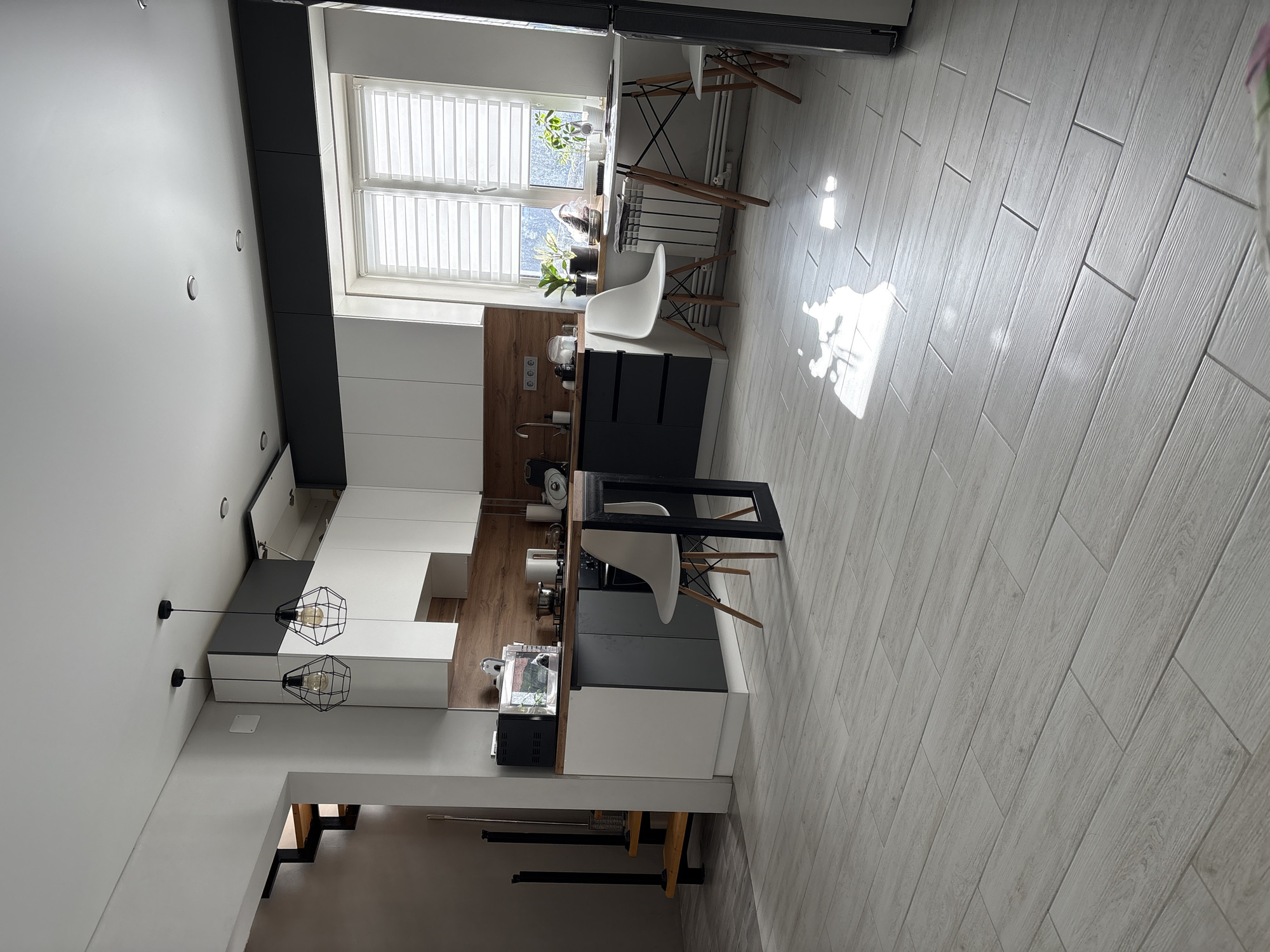 The image displays a modern, well-lit interior kitchen and dining area, stated to be located in the city of Votkinsk, Russia. The floor is covered with light-toned, wood-look plank tiles, reflecting patches of natural sunlight.
The kitchen features white upper cabinets and dark grey lower cabinets and tall units, complemented by a light brown wood-textured backsplash and countertop. Appliances visible on the counter include a black microwave, a silver toaster, an induction stovetop with two pots, and a white kettle. A stainless steel sink with a modern faucet is also present. Two black, geometric cage-style pendant lights hang from the white ceiling over a dark grey kitchen island or bar extension, which is supported by a black metal frame. One white, Eames-style chair with wooden legs is positioned at this bar.
To the right, a large window with white horizontal blinds allows significant natural light, illuminating a small dining area. On the windowsill, several green potted plants are visible. A white, round dining table with wooden legs and black metal supports is placed beneath the window, accompanied by two matching white Eames-style chairs. White plates, bowls, a mug, and a small plant rest on the table. A white radiator is mounted on the wall beneath the window.
On the left side of the image, a minimalist staircase with light wood treads and black metal railings ascends upwards, leading to an upper level. The walls throughout the space are predominantly white or light grey, with several recessed ceiling lights also present. No individuals are visible in the scene.