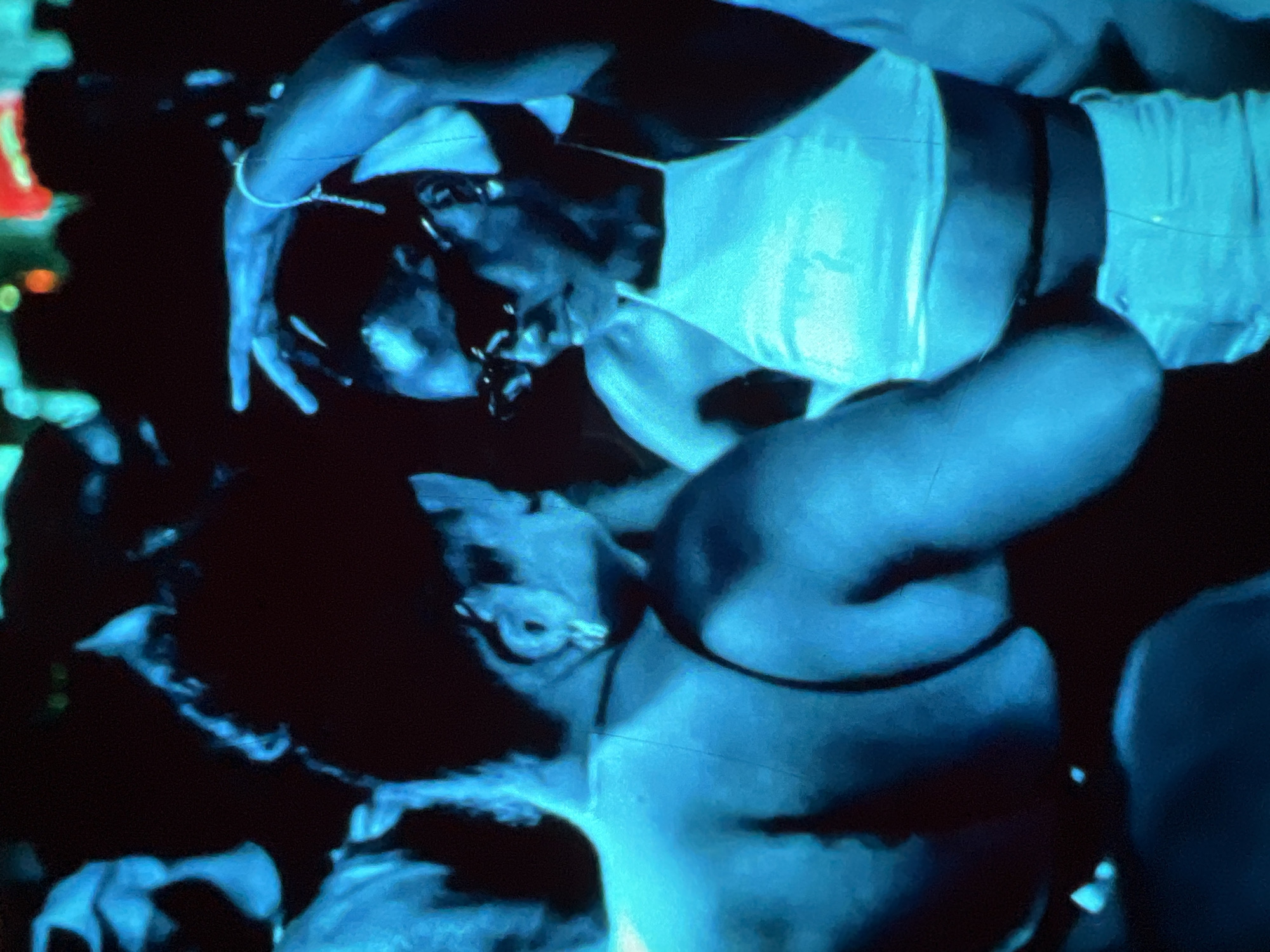 This is a medium shot, taken at night in a dimly lit indoor venue in Lagos, Nigeria. The scene appears to be a crowded event, possibly a party or club, with an intense blue and green lighting effect. Two individuals are the primary subjects, both appearing to be of African descent and dressed in attire suitable for a social gathering.
In the foreground, on the left, a person with dark hair pulled back is seen from the back and side. They have broad shoulders and are illuminated by the blue light, revealing their back and right arm. Their skin has a blueish hue due to the lighting.
To their right and slightly behind is another individual, facing left. This person is wearing sunglasses and a light-colored top, possibly a halter top or a sleeveless shirt. Their left arm is raised, with a bracelet on their wrist, and their fingers are extended. Their facial features are partially obscured by the sunglasses and the lighting, but their profile is visible. The lighting on this individual is a bright turquoise, highlighting their upper body and face.
The background is dark and indistinct, with a crowd of people blurred and out of focus. Some faint colored lights, possibly from stage lighting or other patrons, are visible in the upper part of the image, suggesting a dynamic and possibly energetic environment. The overall impression is one of a vibrant, active social scene in Lagos at night, characterized by strong, artificial lighting.