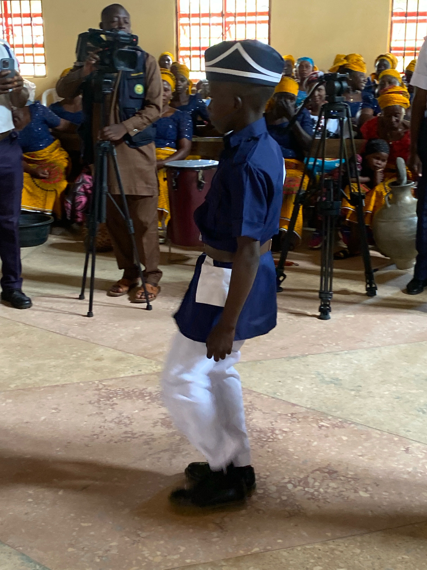 A young boy dressed in a dark blue uniform with white pants and a white cap with blue trim, is standing in the foreground facing right. The boy is wearing black shoes. Behind him, a man in a brown shirt and vest is operating a video camera on a tripod. In the background, a group of people, mostly women wearing yellow headscarves and blue and yellow attire, are seated. A large clay pot is visible to the right of the frame. The environment appears to be indoors with tiled floors and windows with bars. The scene is likely taking place in Ohit, Nigeria, possibly during a ceremony or event.